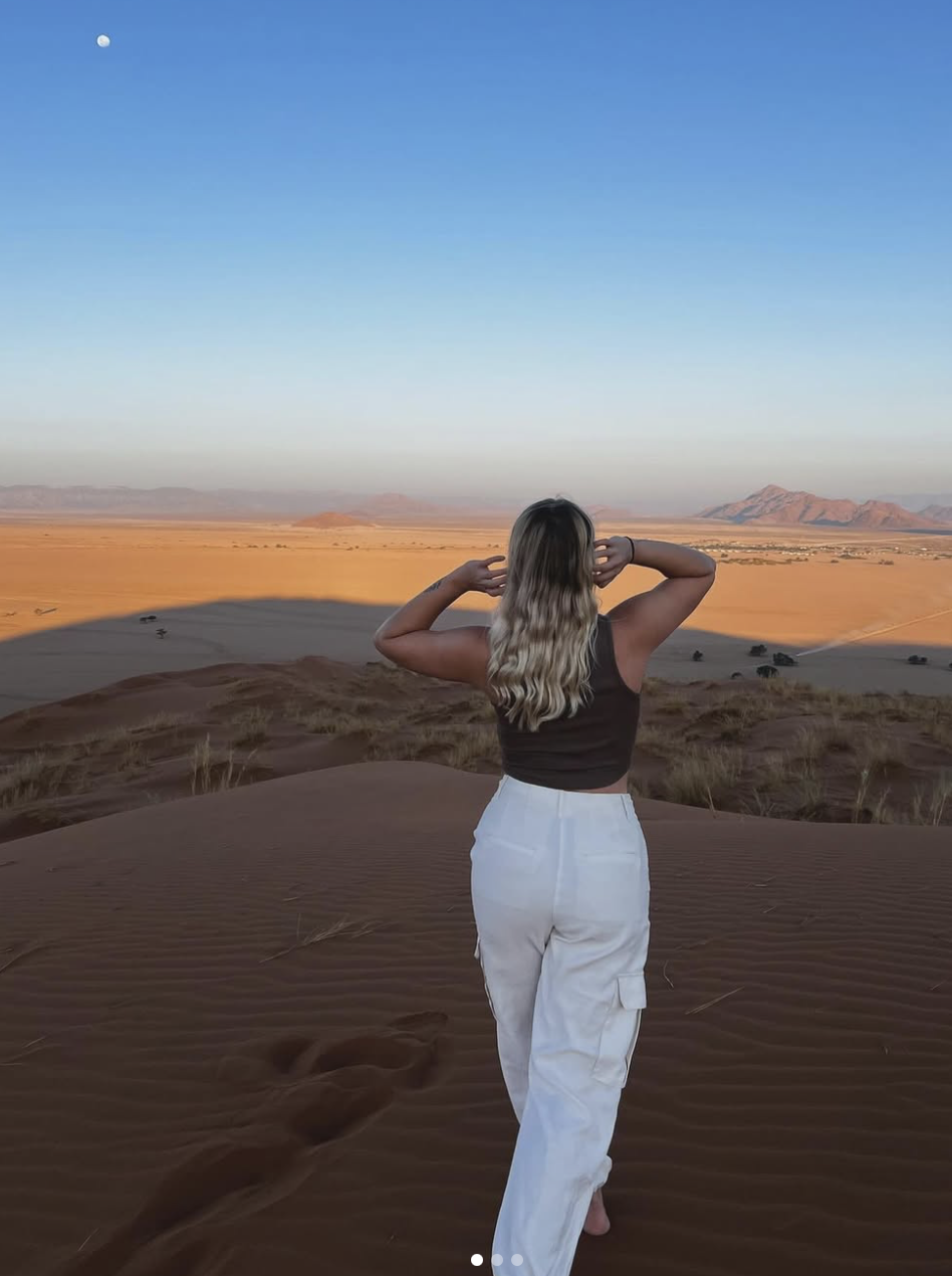 Une femme marche dans un désert de sable en regardant un coucher de soleil ou un lever, avec un ciel bleu clair et la lune visible dans le ciel.