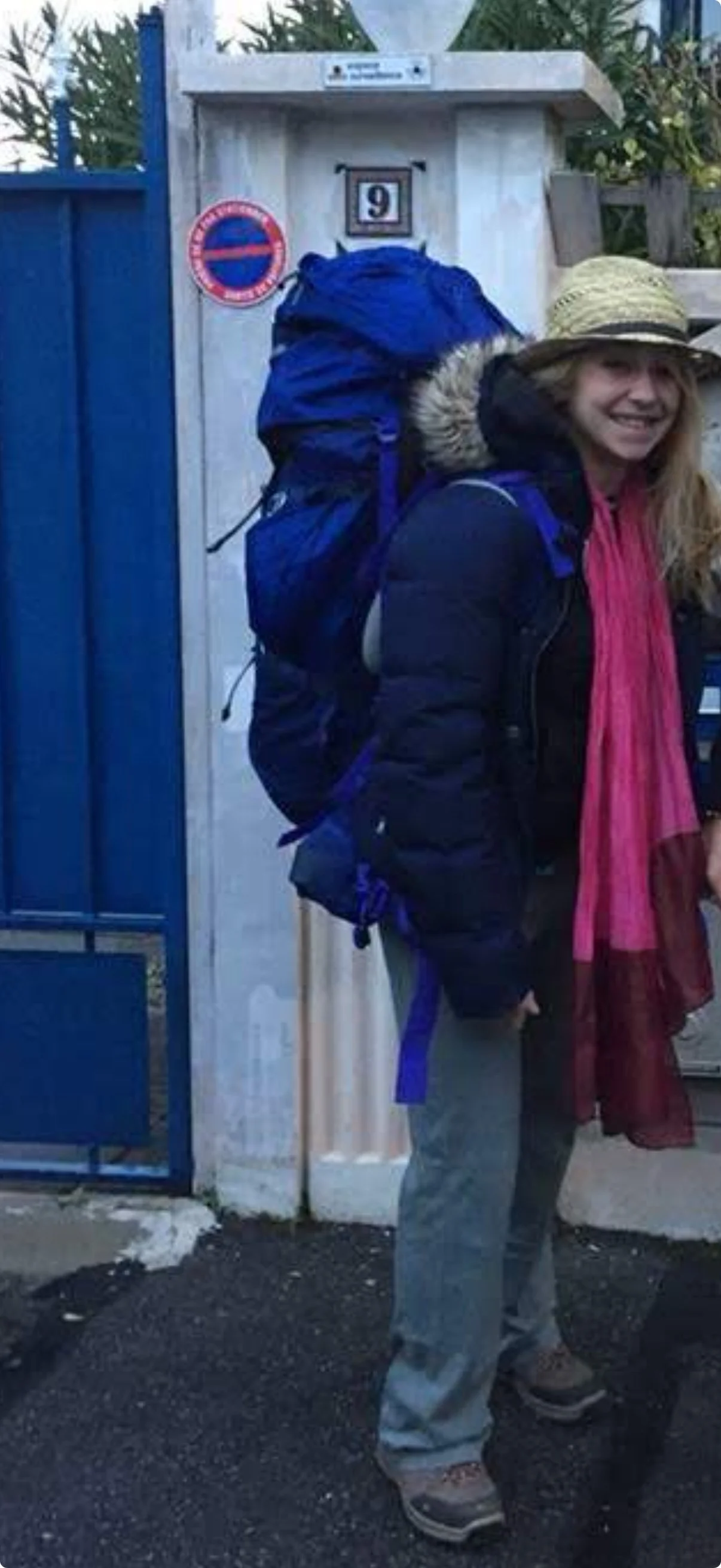 Une femme souriante portant un sac à dos bleu, un chapeau en paille et une veste noire, se tenant devant une porte bleue à côté d'un mur en pierre.