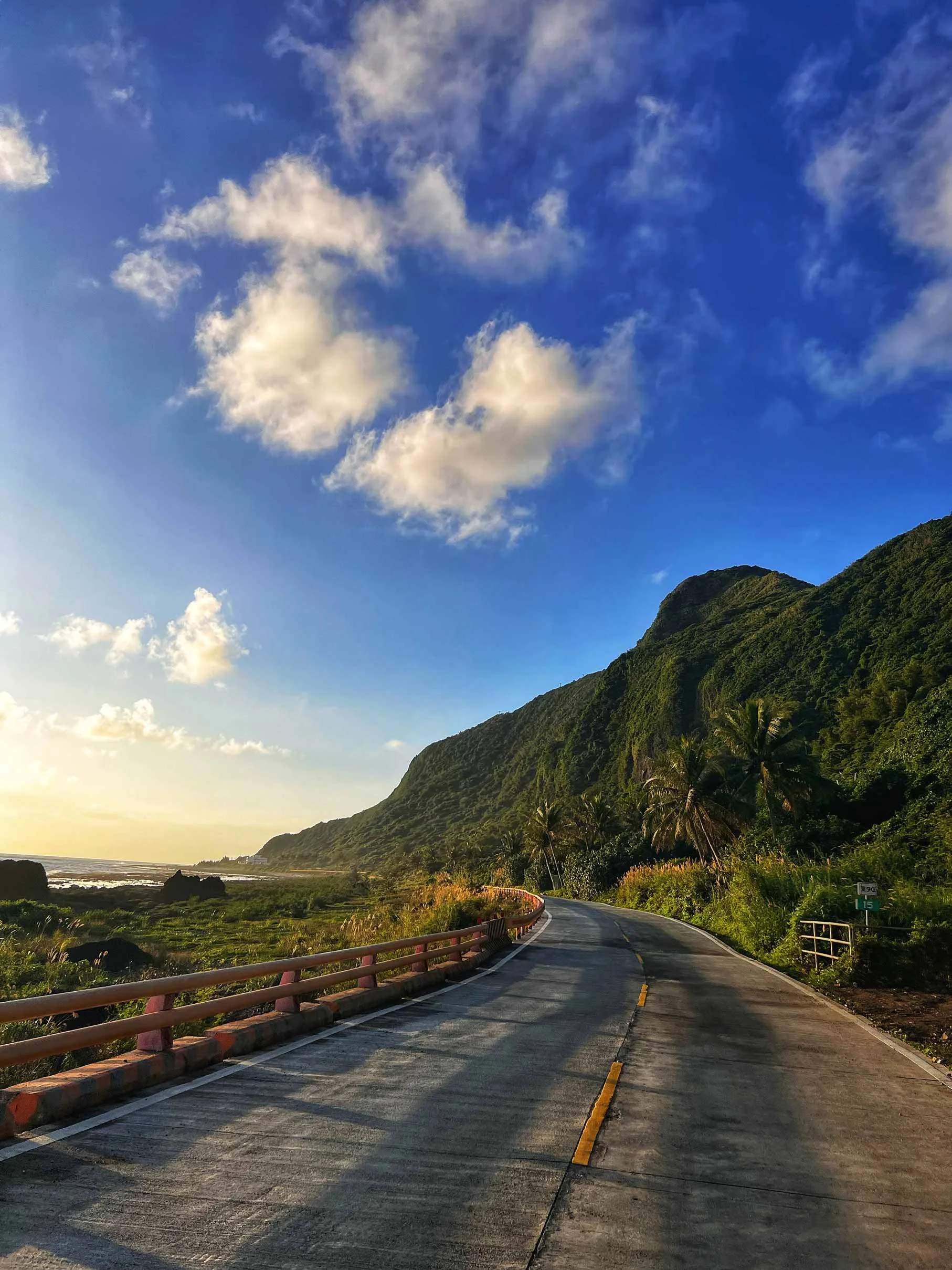 Route sinueuse en bord de mer entourée de montagnes verdoyantes sous un ciel bleu avec quelques nuages