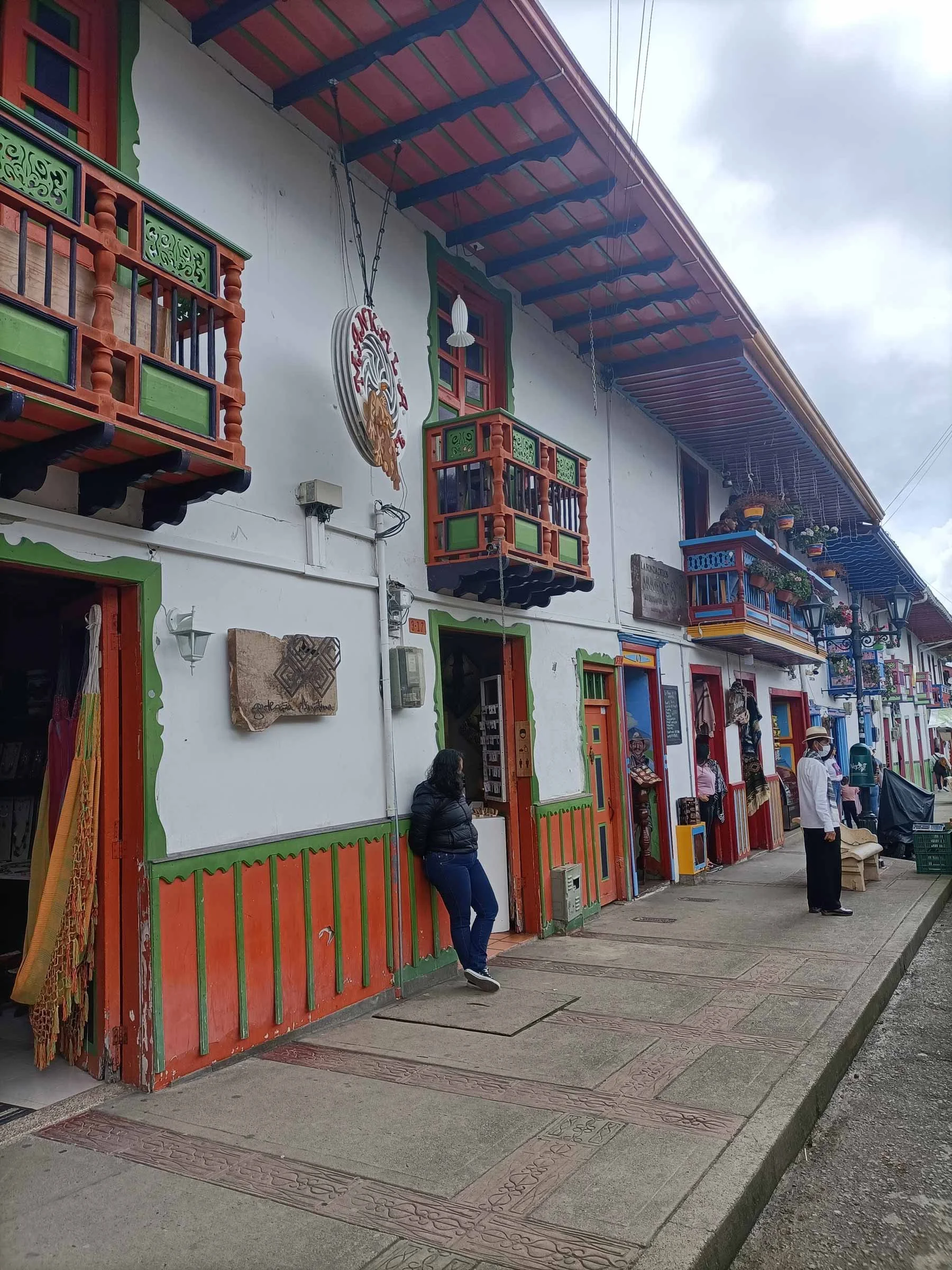 Rue avec des bâtiments colorés, balcons en bois, boutiques ouvertes, personnes marchant, ciel nuageux.