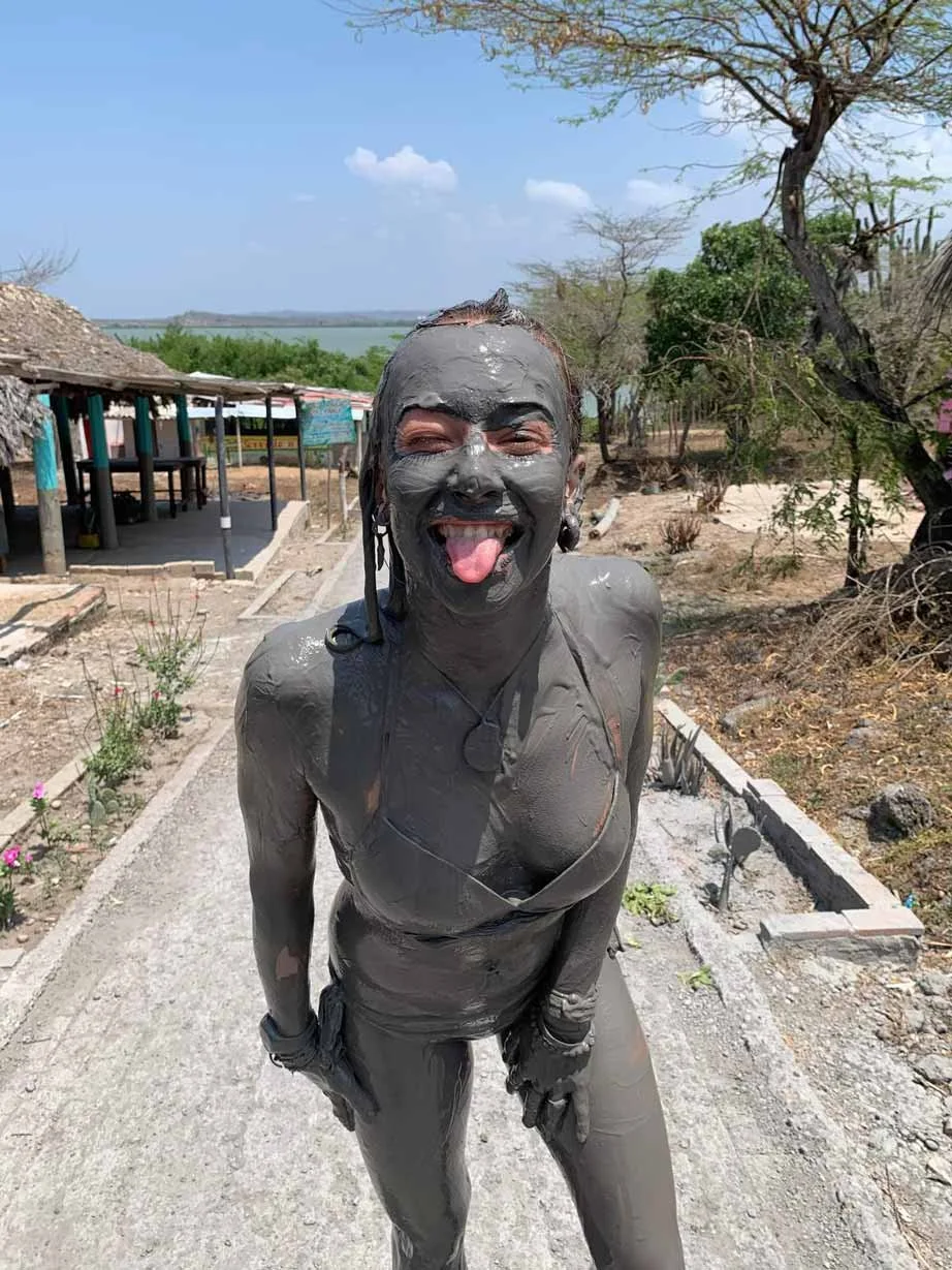 Charlotte couverte de boue tirant la langue, située en plein air avec des arbres, un ciel bleu et une structure en fond.