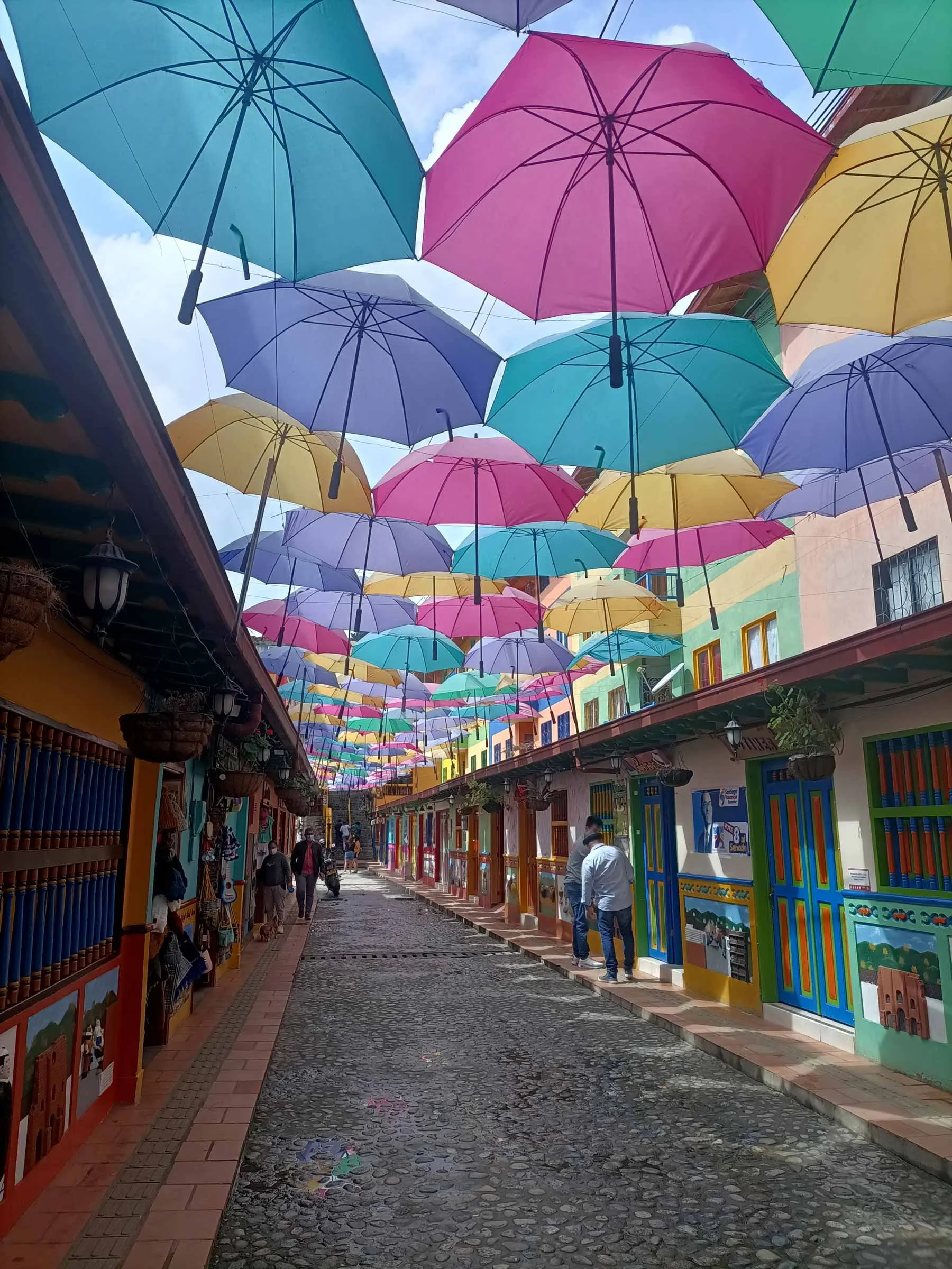 Rue pavée bordée de maisons colorées avec des parapluies suspendus au-dessus, créant un plafond artistique. Quelques personnes marchent ou discutent.