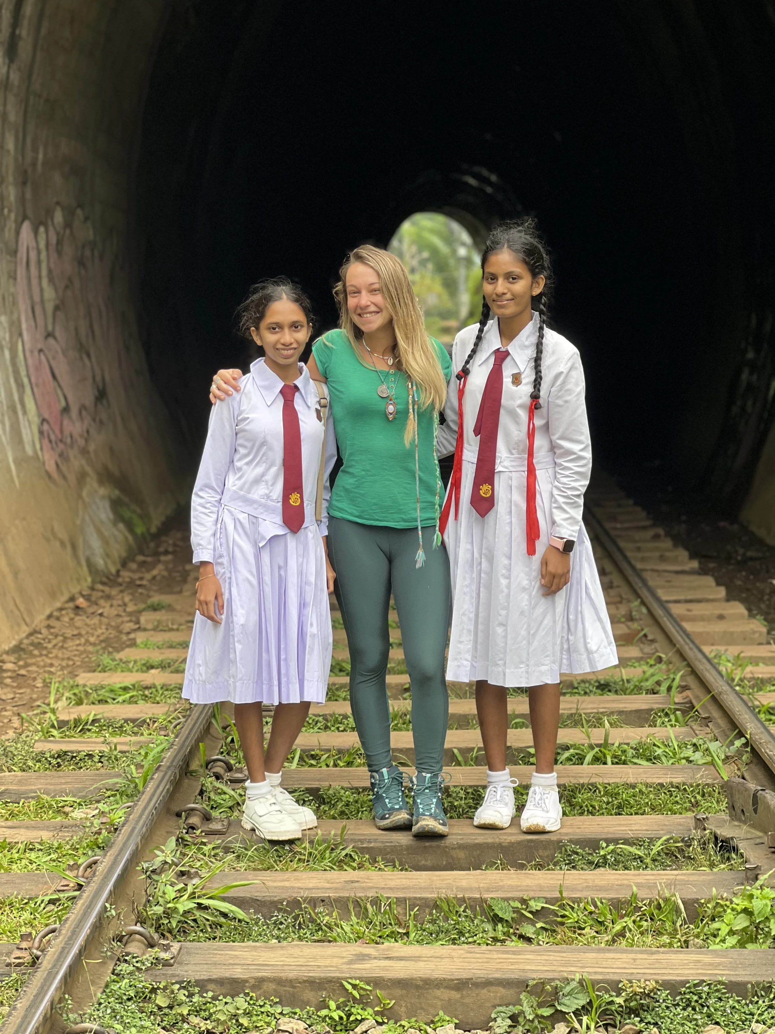 Trois femmes debout sur des voies ferrées dans un tunnel sombre, deux en uniformes scolaires blancs avec cravates rouges, et une femme en vêtements décontractés verts, souriantes ensemble.