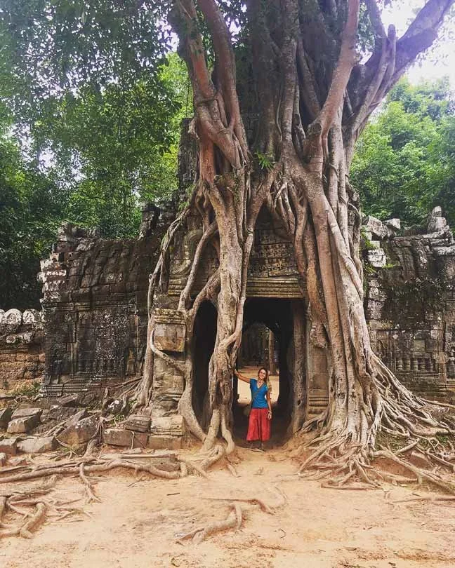 Une femme debout sous une vieille structure en pierre envahie par de grands arbres aux racines torsadées, entourée de végétation dense.