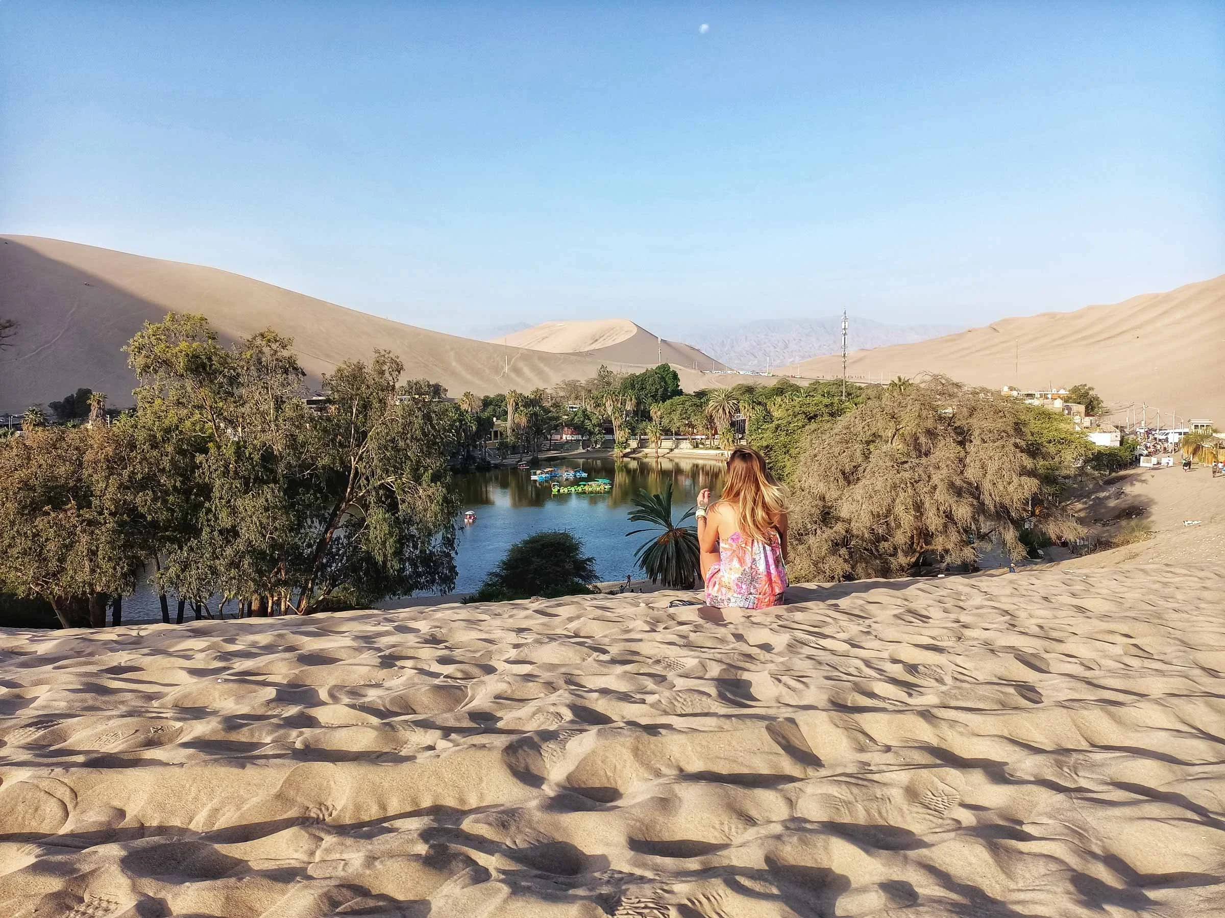Une femme assise dans le sable du désert, regardant un oasis avec des arbres, une rivière, et des dunes de sable au loin.