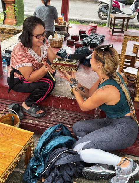 Une femme vendant des objets artisanaux à une cliente assise par terre, dans un stand de marché en plein air, avec plusieurs articles décoratifs autour d'elles.
