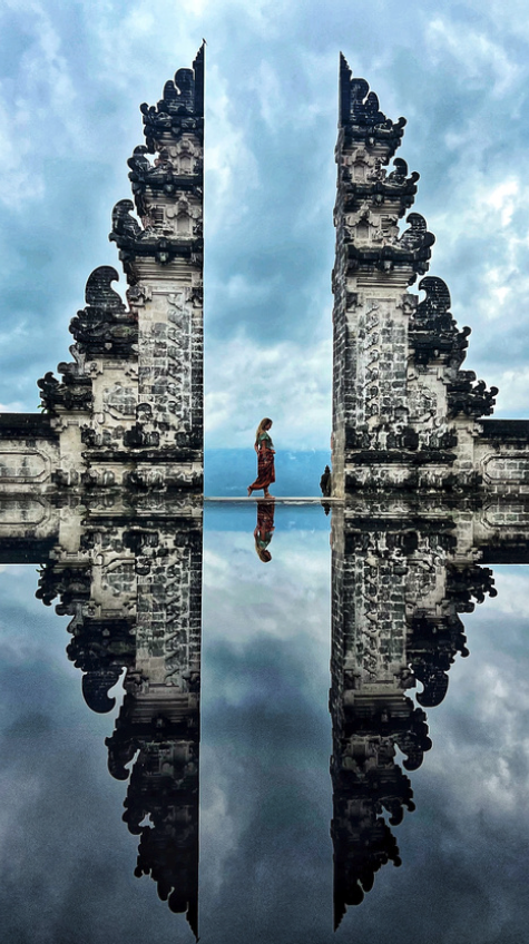 Femme en robe flottante se tenant entre deux structures de pierre ornées, avec leur reflet dans un miroir d'eau, ciel nuageux en arrière-plan.
