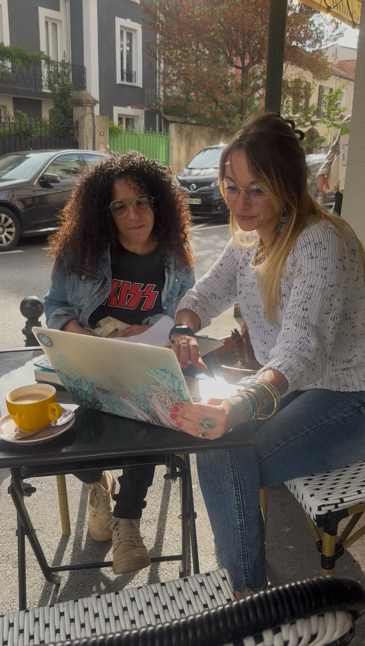 Deux femmes regardant un ordinateur portable à une table de café en terrasse, avec une tasse de café jaune sur la table, dans une rue urbaine avec des voitures et des bâtiments en arrière-plan.