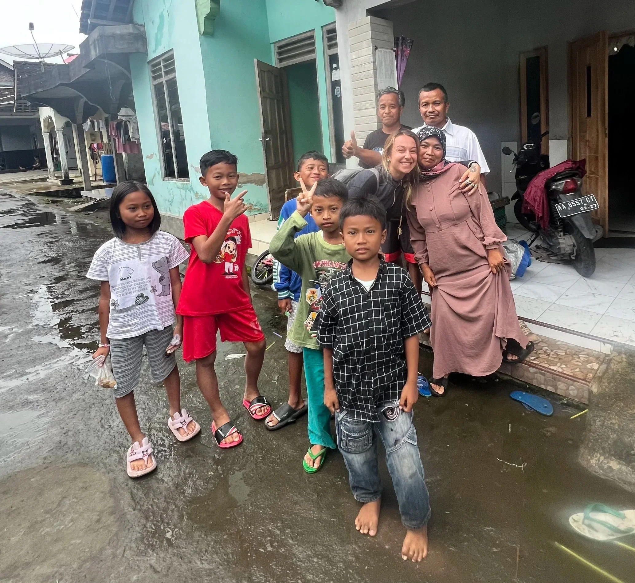 Groupe d'enfants et d'adultes devant une maison dans une rue mouillée, certains faisant des signes de paix ou de victoire, avec un chien et une moto en arrière-plan.