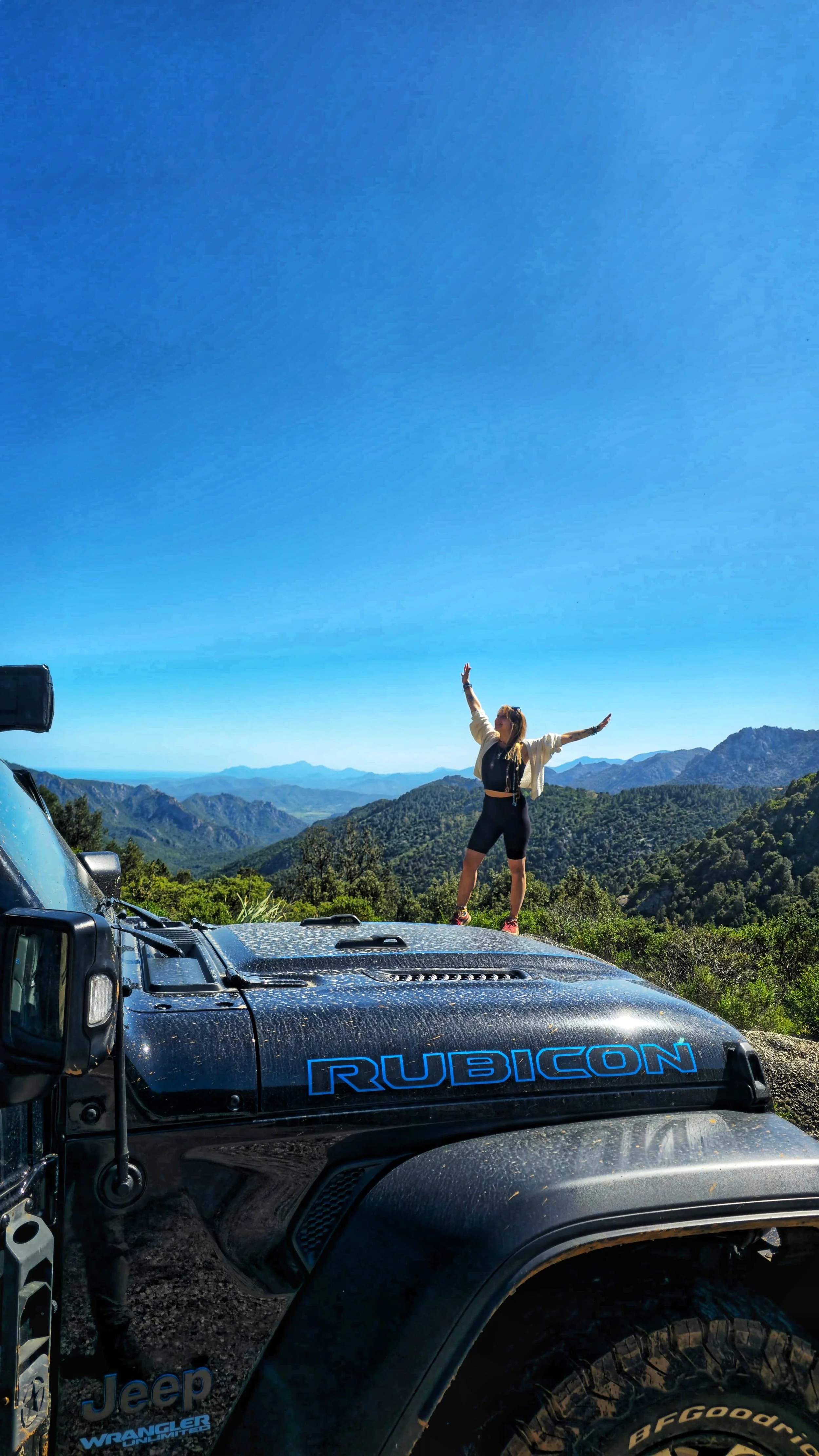 Une femme se tient sur le capot d'un Jeep Rubicon, dans un paysage de montagnes verdoyantes sous un ciel bleu clair.