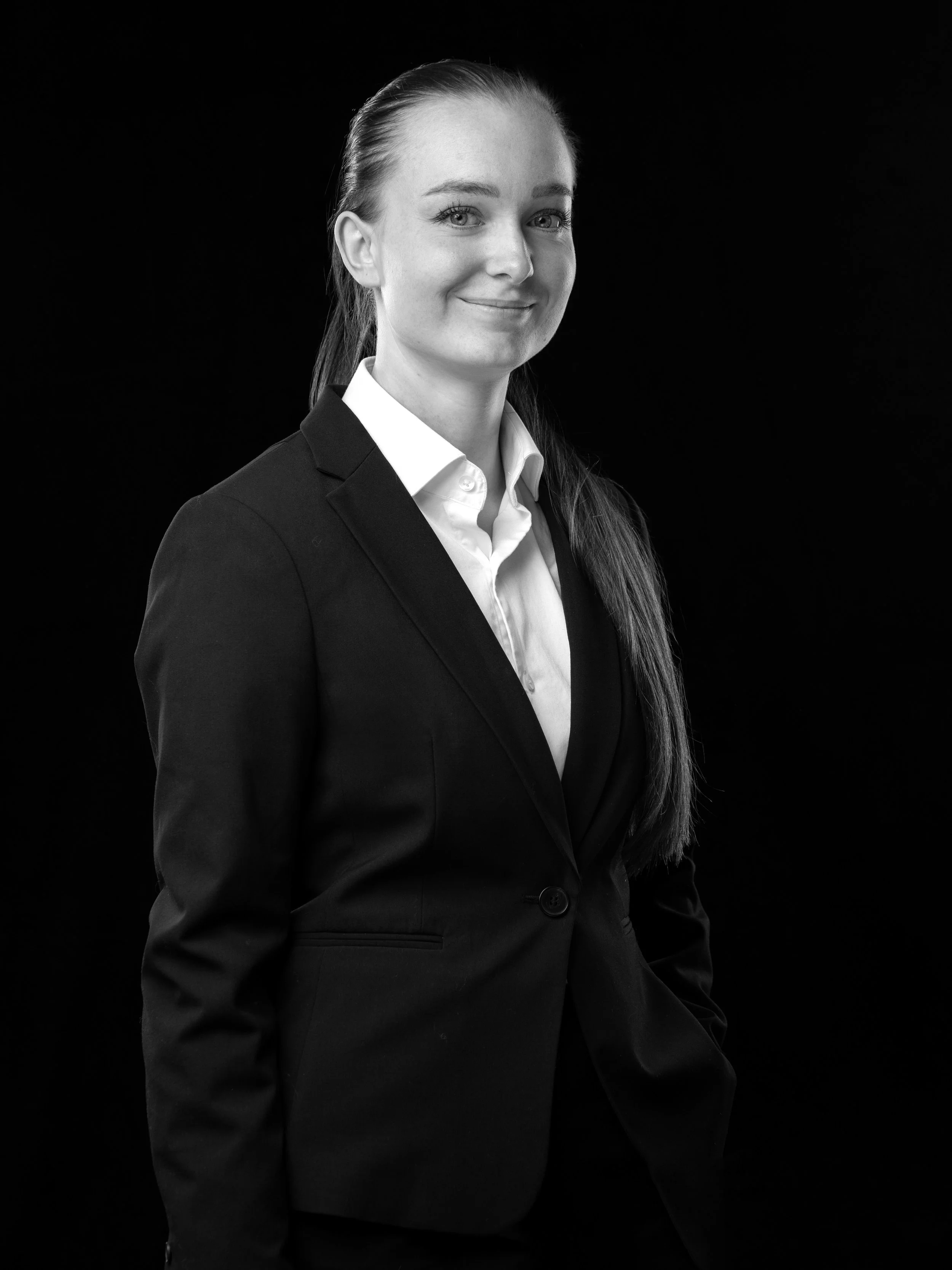 En mann smiler, iført en lys skjorte og svart blazer, i svart-hvitt fotografi mot en svart bakgrunn.