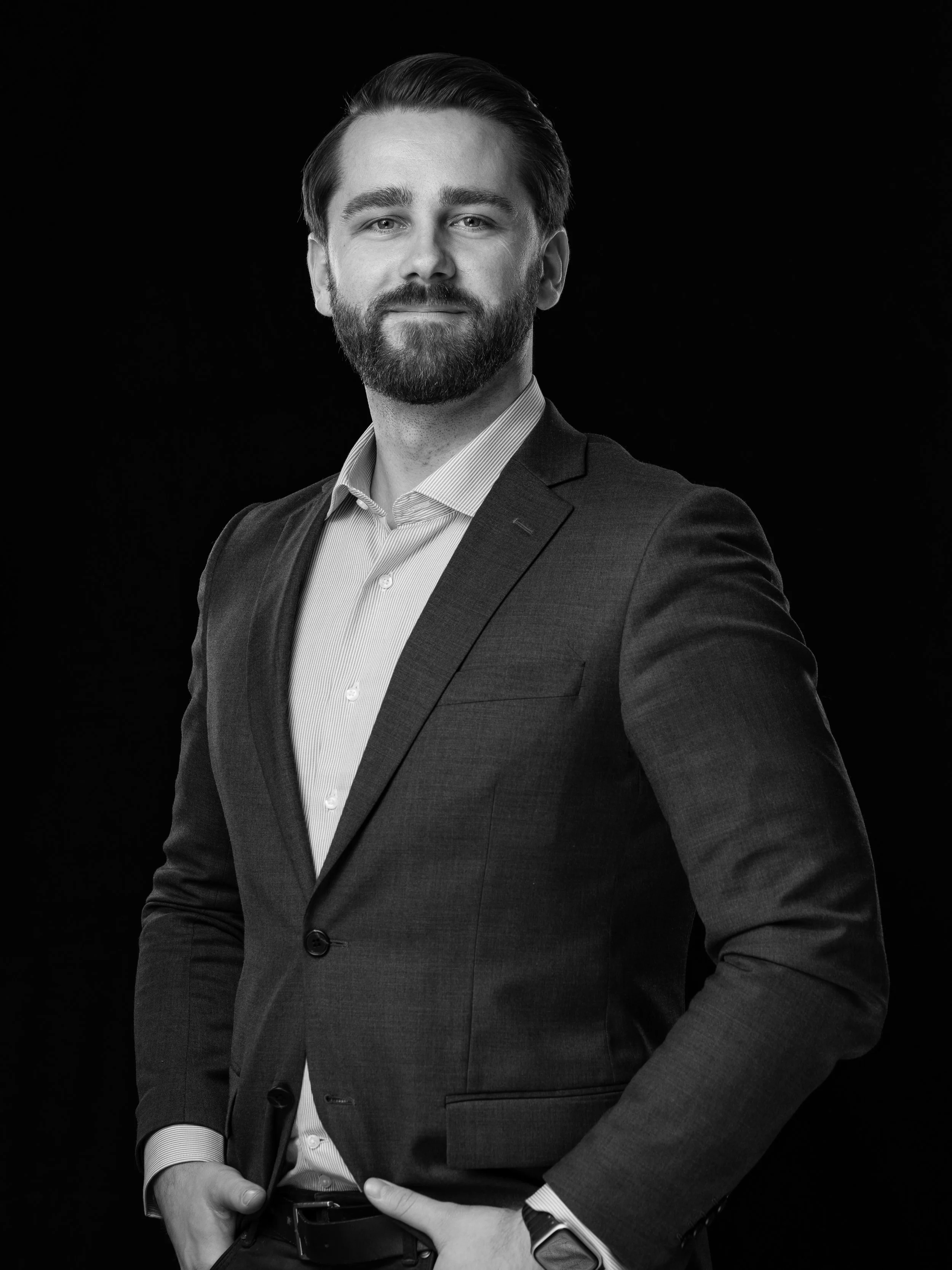 Svart-hvitt bilde av en smilende mann i dress og hvit skjorte mot svart bakgrunn.