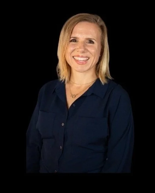 A smiling woman with shoulder-length blond hair, wearing a dark blue button-up shirt and gold necklace, against a black background.