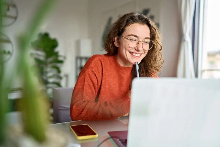 Young woman with curly hair and glasses smiling while using a laptop and drinking from a straw, sitting at a desk in a bright room.