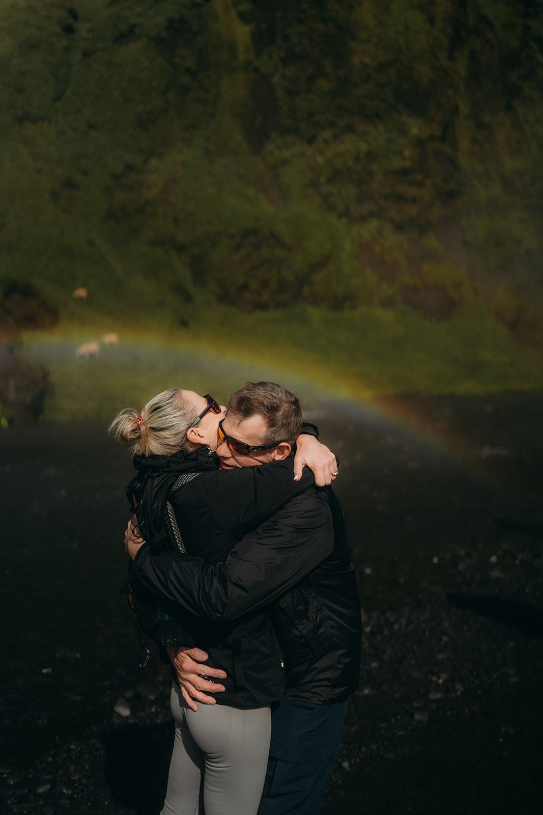 Pár se drží v objetí u vodopádu Skogafoss na Islandu. Za nimi je vidět duha. Autentická chvíle plná lásky a přírody.