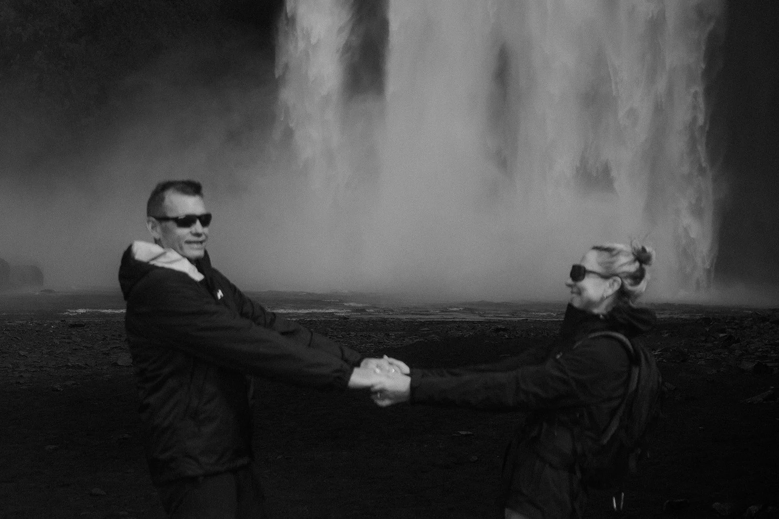 černobílá fotografie muže a ženy na dovolené na Islandu. Pár se drží za ruce před vodopádem Skogafoss. Zaostřeno je na vodopád v pozadí. Pár v popředí je rozostřený.