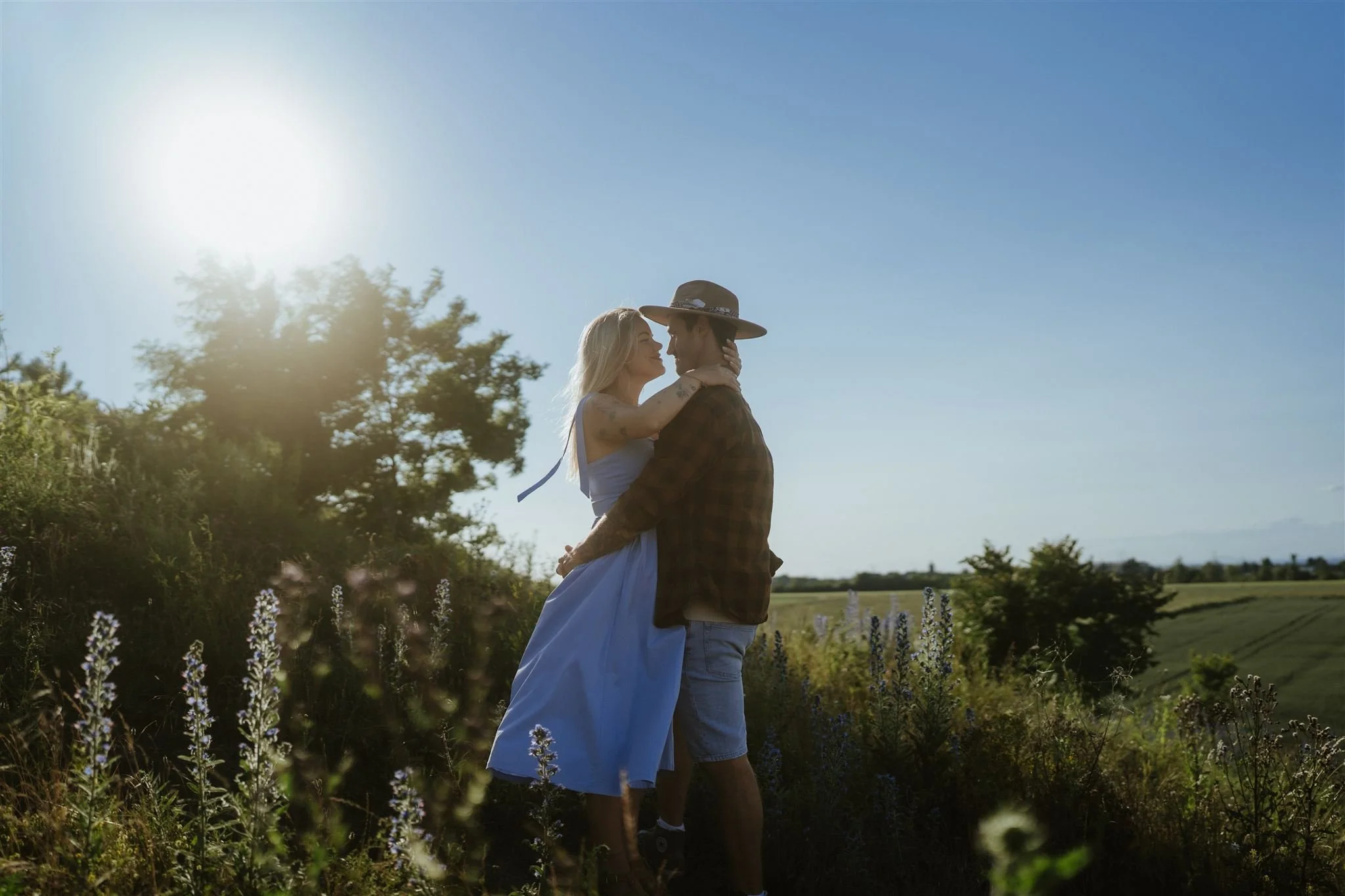 Pár v objetí, v přírodě při západu slunce. Žena má modré šaty, dívá se na muže.