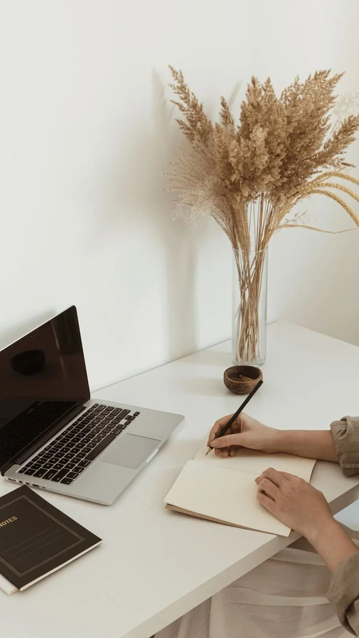 A woman writing in a notebook next to an open laptop.