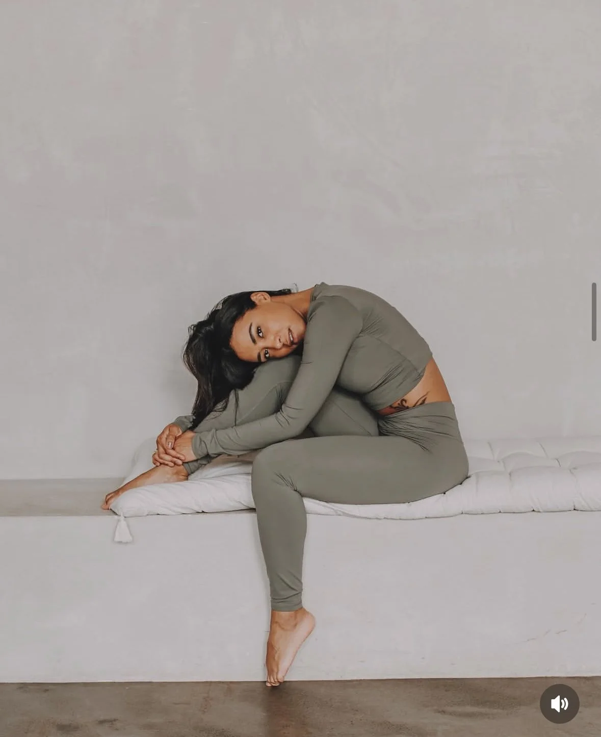 Woman in gray athletic wear sitting cross-legged on a cushioned bench against a light gray wall.