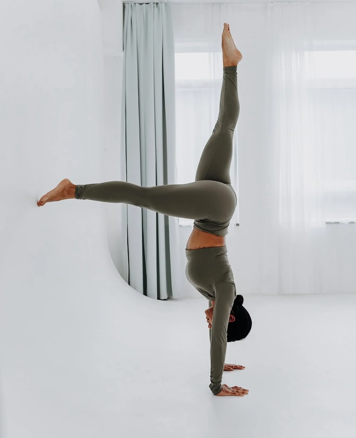 Person performing a handstand in a minimalist white room with curtains, wearing green activewear.