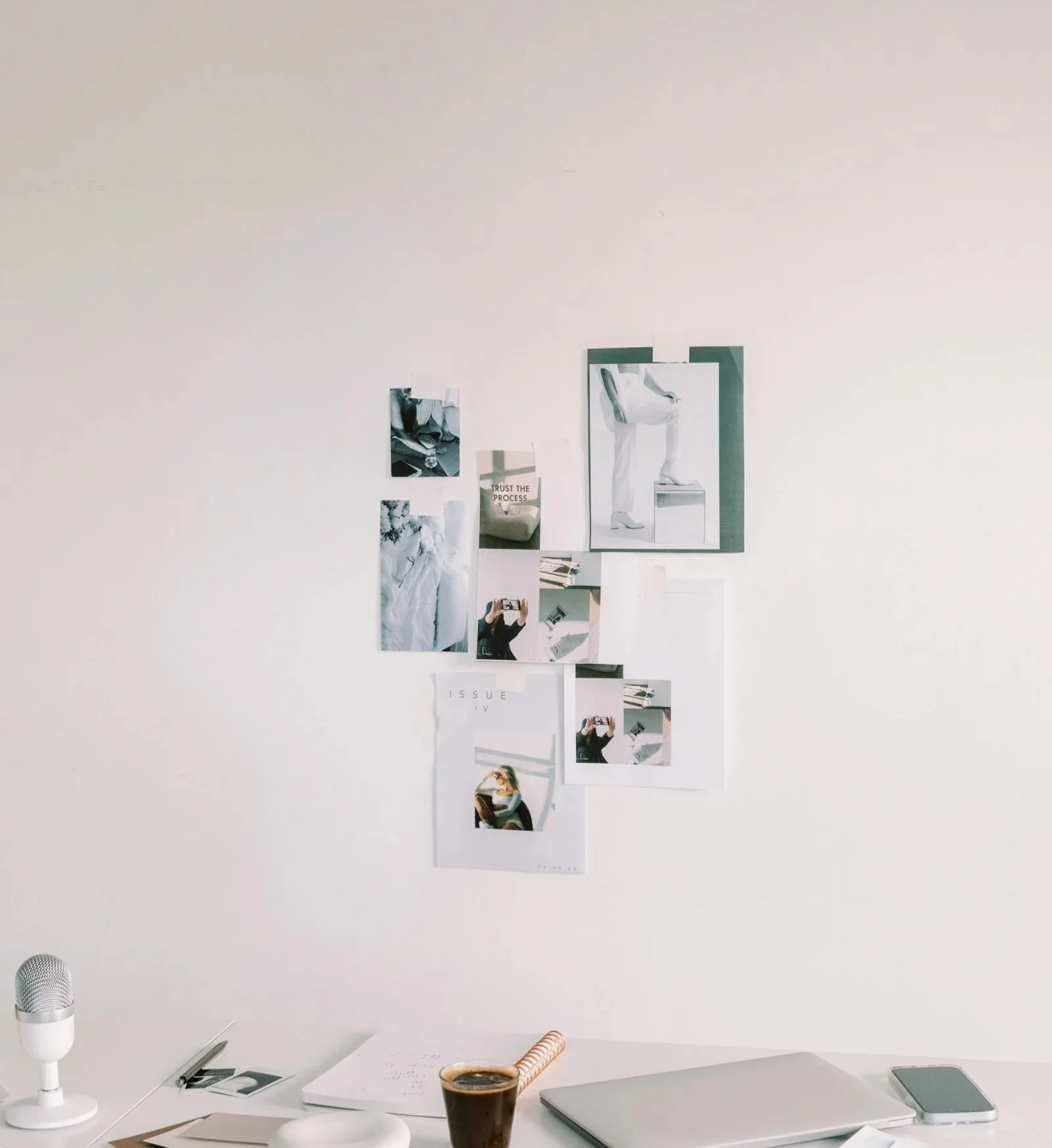 Photo of a minimalist workspace with a white desk, a microphone on the left, a coffee cup in the center, a closed laptop, a phone, notebooks, and photos of a person on the wall.