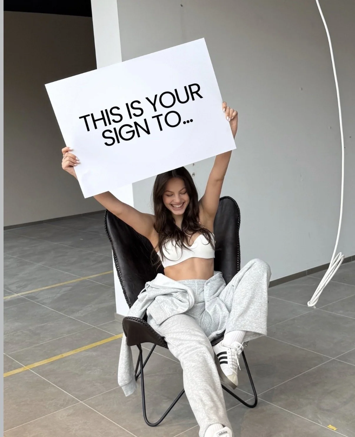 A young woman sitting in a black chair indoors, holding a large white sign that reads 'This is your sign to...' and smiling with her eyes closed. She is wearing a white sports bra, gray sweatpants, and white Adidas sneakers, with a gray sweatshirt draped over her lap.