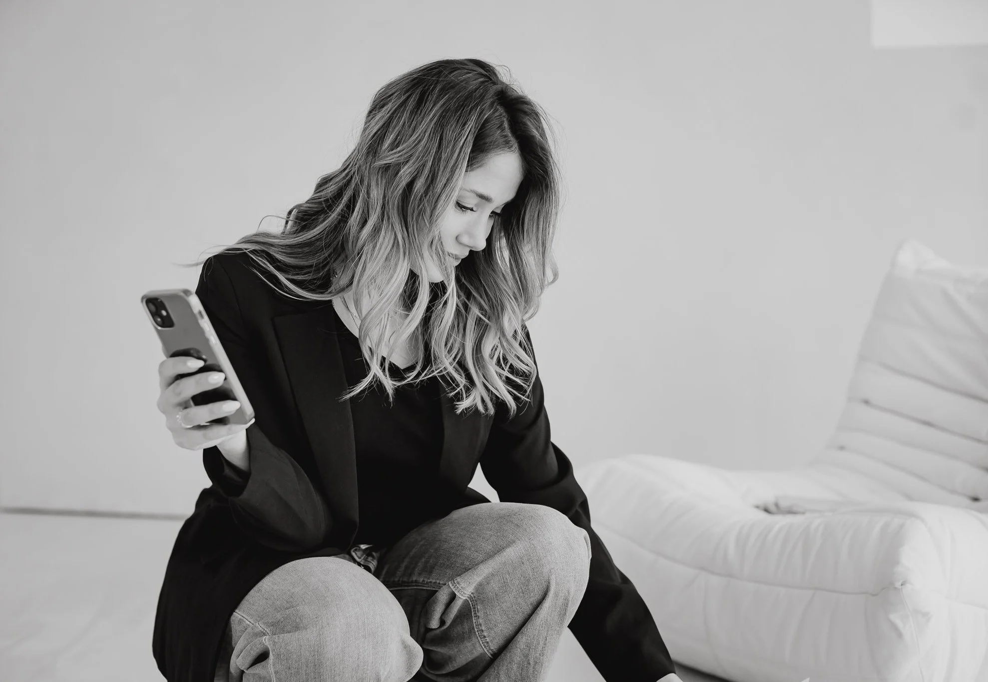 Black and white image of a woman with long hair holding a smartphone, sitting on white furniture.