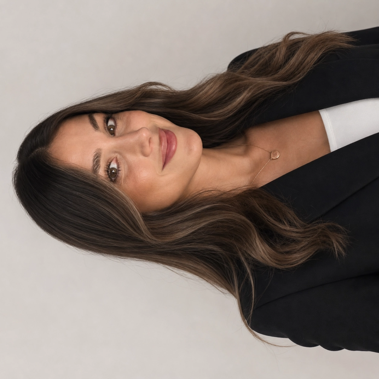 A woman with long wavy brown hair, wearing a black blazer, white top, and a delicate necklace, looking into the camera with a slight smile.