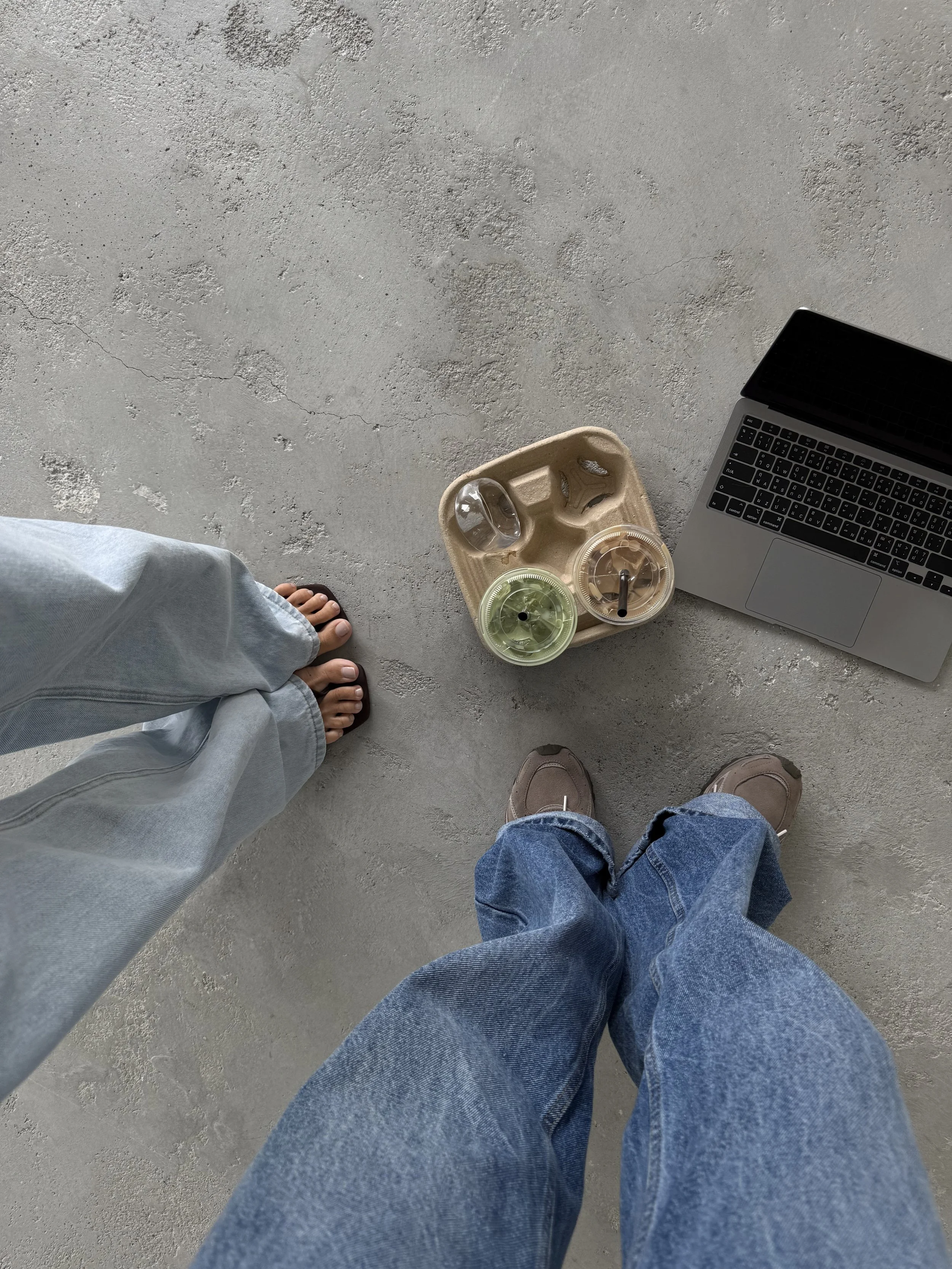 From a top-down perspective, two people are standing on a concrete floor while wearing jeans and sneakers. One person has beige sneakers, and the other has brown sneakers. There is a tray with three drinks and a laptop nearby.