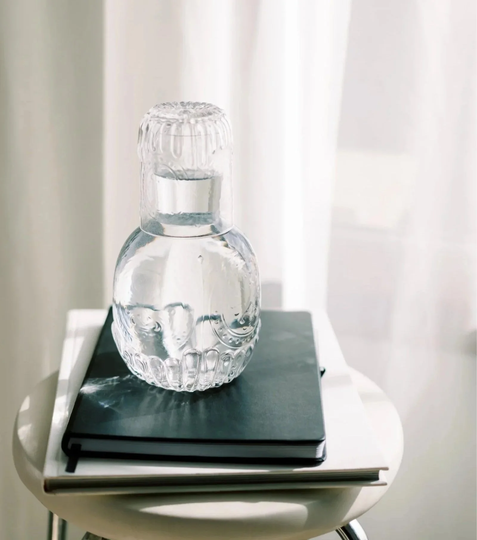 A clear glass skull-shaped bottle filled with water, placed on a black closed notebook on a white surface with a white background.