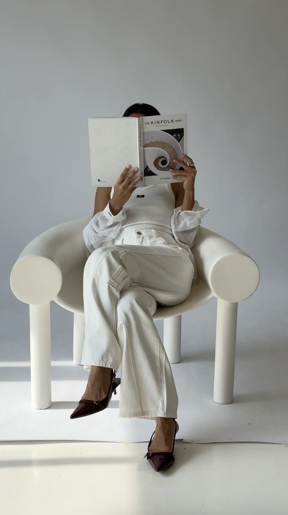 A woman sitting on a cream-colored modern chair, reading a magazine titled 'The Kinfolk Home'. She is dressed in white clothing with dark high heels, and her face is hidden behind the magazine, against a plain light gray background.