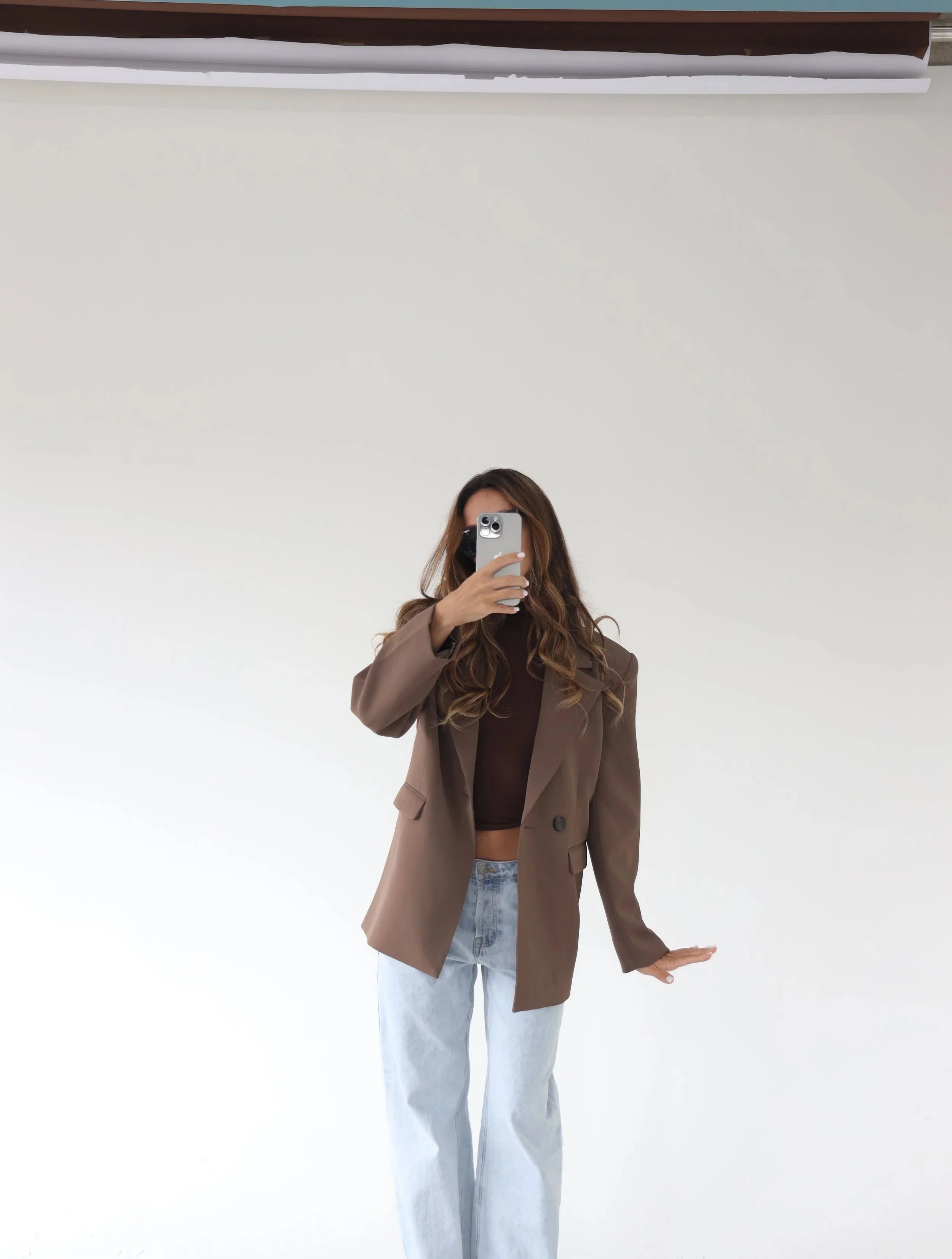 A woman with long hair taking a mirror selfie with a smartphone in a minimalistic space with white walls, wearing a brown blazer, black top, and light blue jeans.