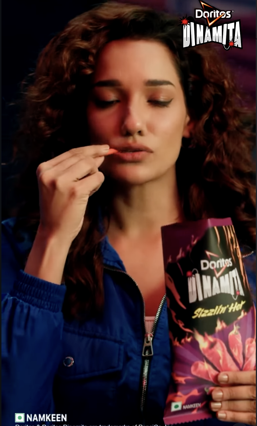 A woman with curly brown hair holding a bag of Doritos Dynamite Stezillit Hot chips, standing against a dark background with the Doritos Dynamite logo in the top right corner.