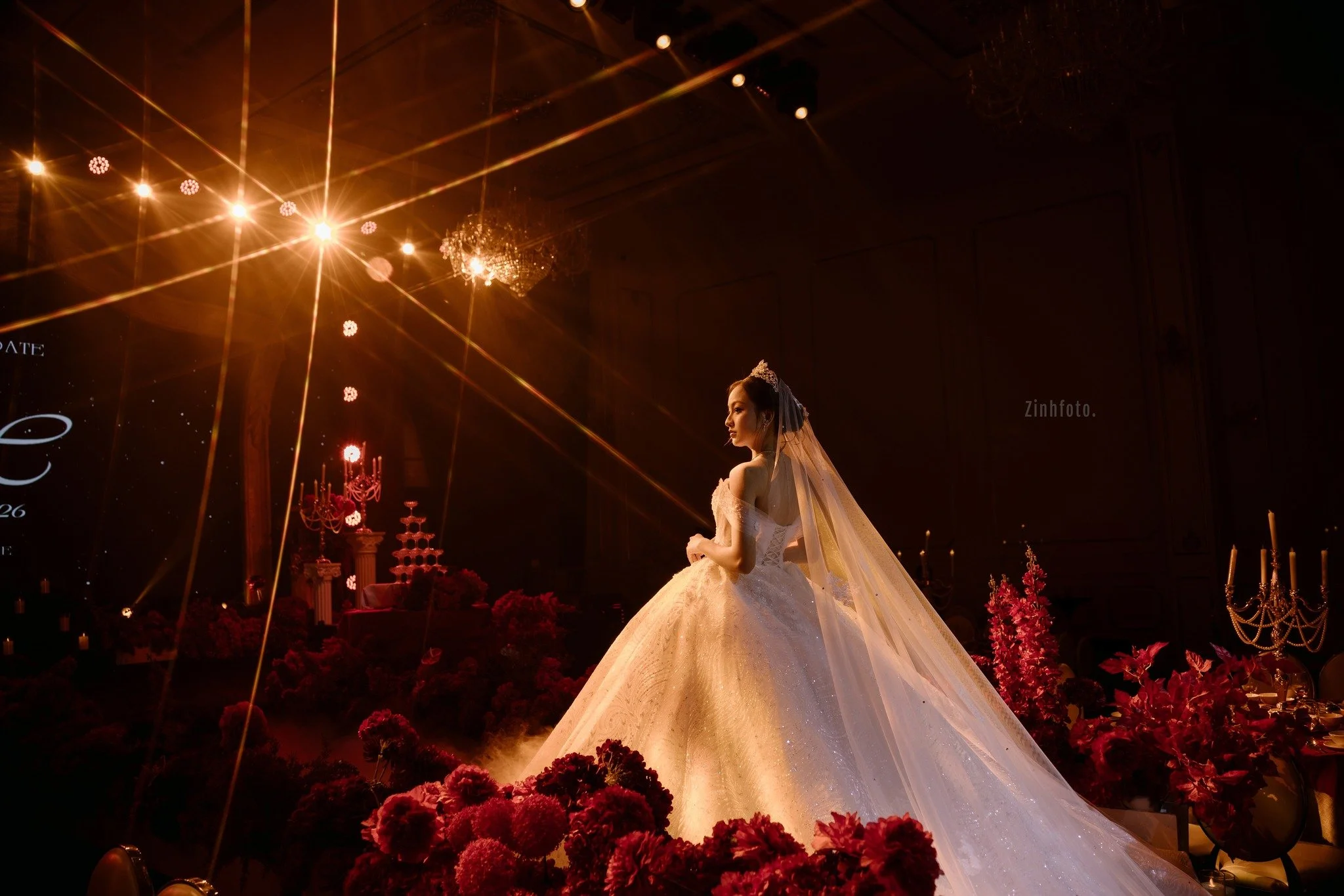 A bride in a white wedding dress with a long train and veil stands in a dimly lit room decorated with pink flowers and candelabras, with bright overhead lighting creating a starburst effect.