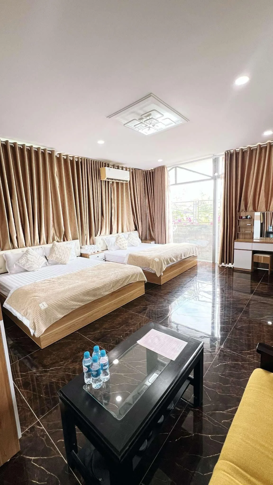 A hotel room with two beds, brown curtains, a dark brown marble floor, a small table with bottled water, and a balcony with glass door.