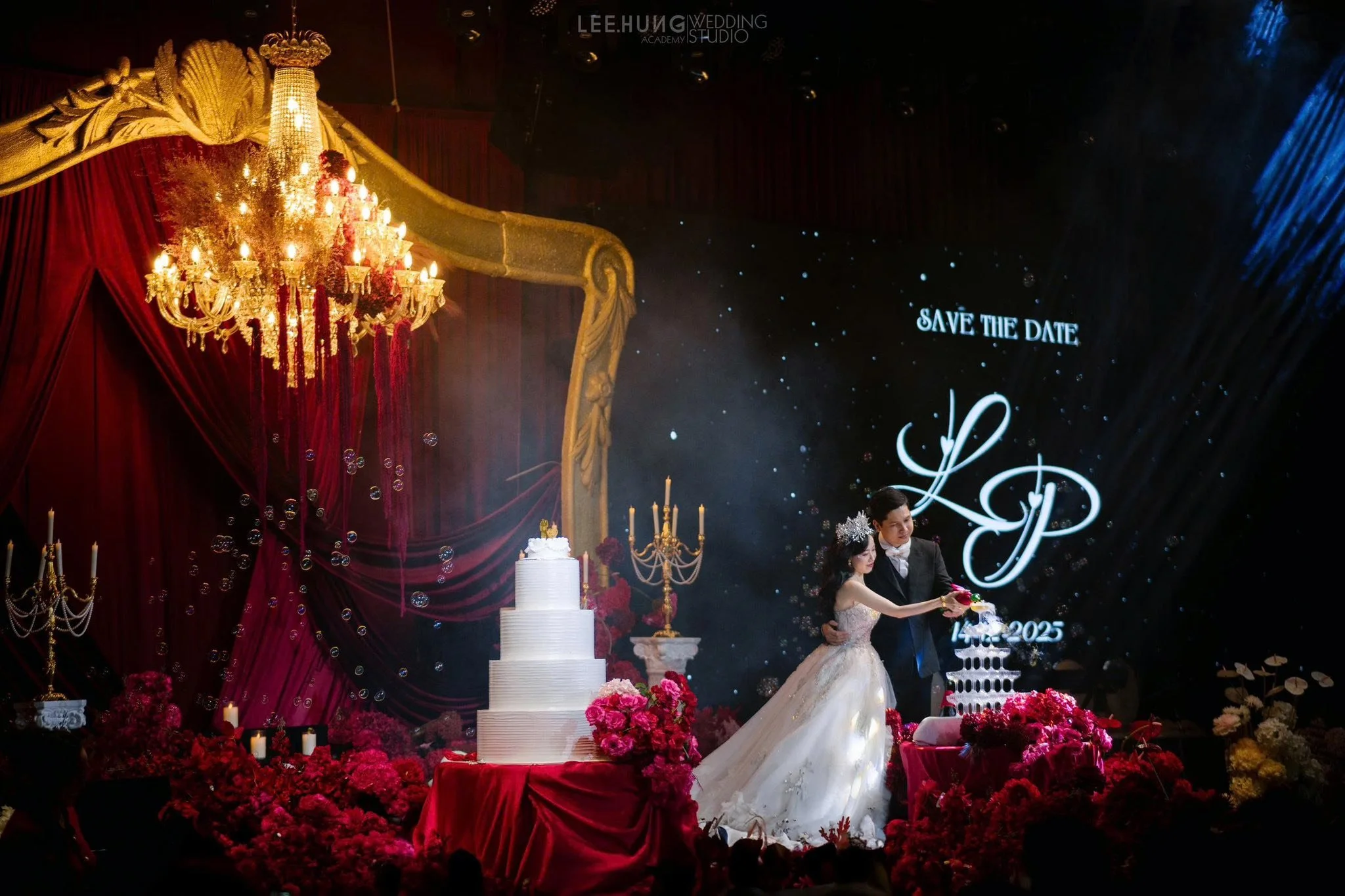 Bride and groom cutting wedding cake on decorated stage with red drapery, chandelier, flowers, and a large screen that displays祝SAVE THE DATE and initials with wedding date.