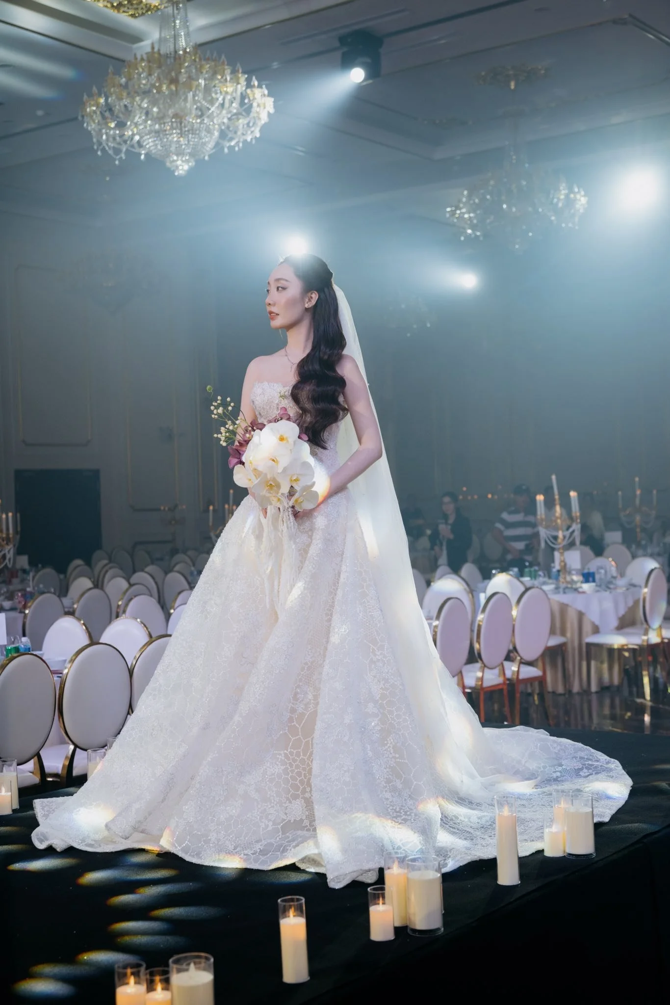 A bride in a wedding dress holding a bouquet of flowers standing in a decorated banquet hall with chandeliers and candles.