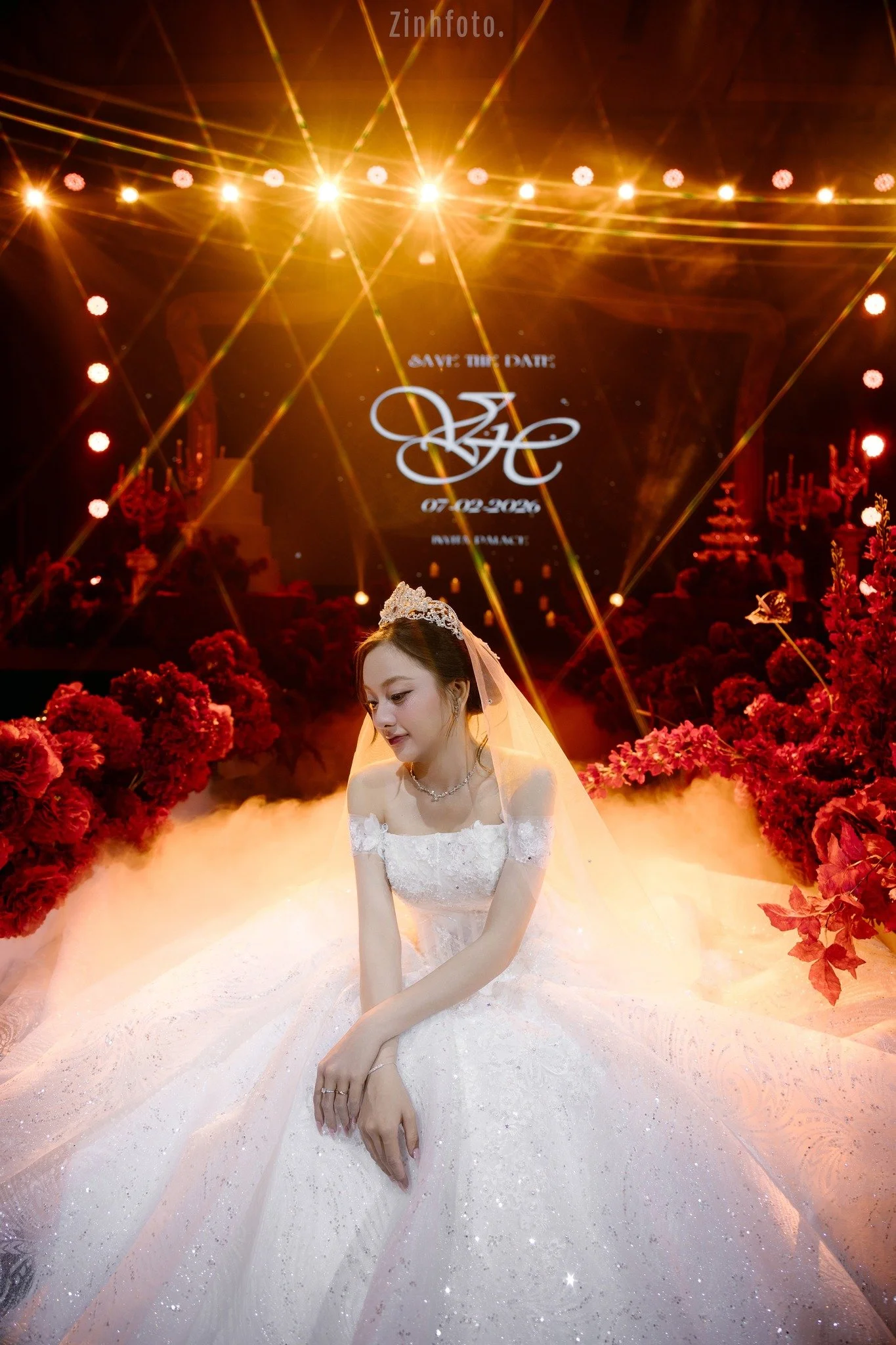 A bride in a white wedding gown, wearing a tiara and veil, sitting on the floor surrounded by red floral arrangements, with a bright stage and laser lights in the background.