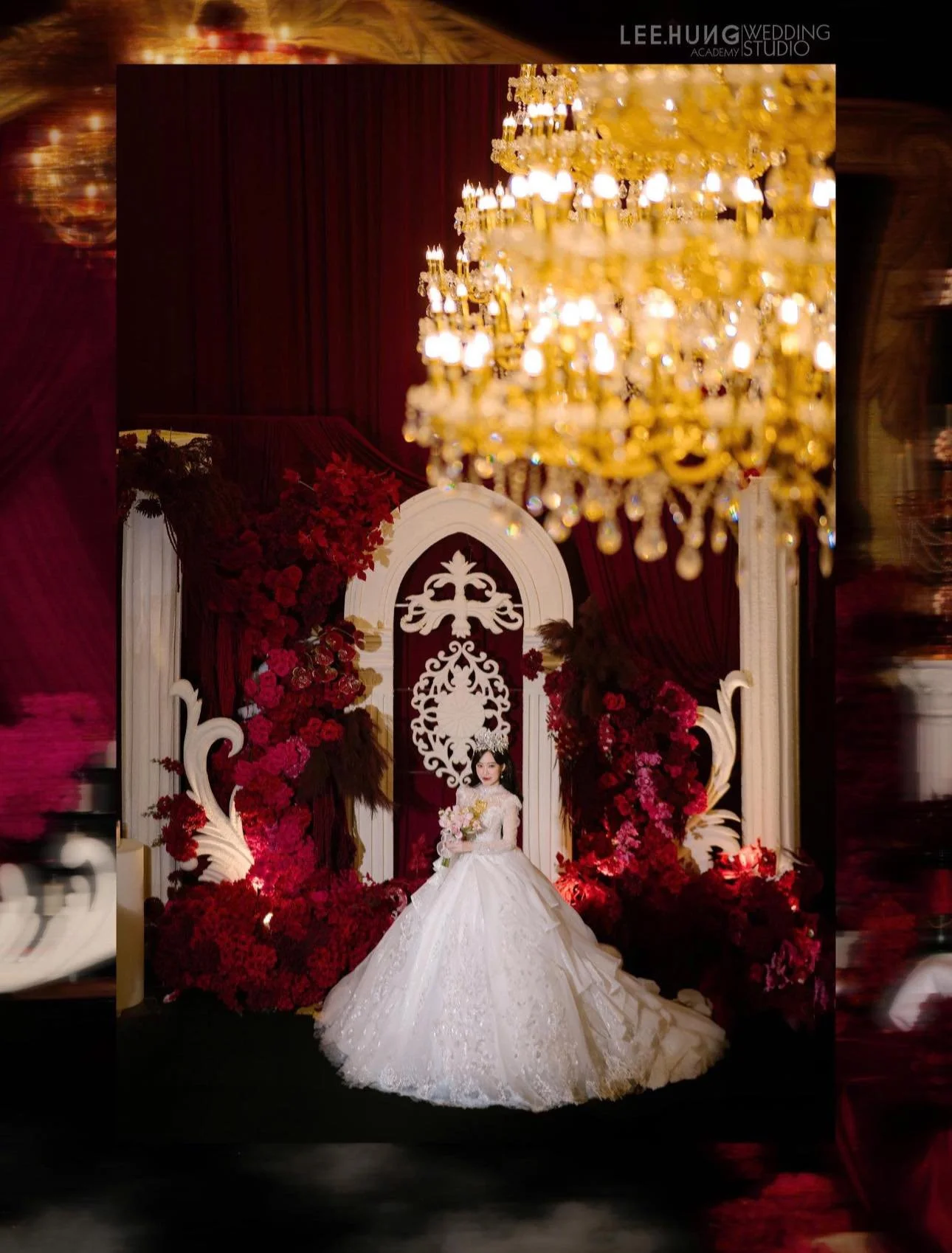 A bride in a white wedding gown holding a bouquet stands in front of a decorative backdrop with red floral arrangements and a chandelier overhead.