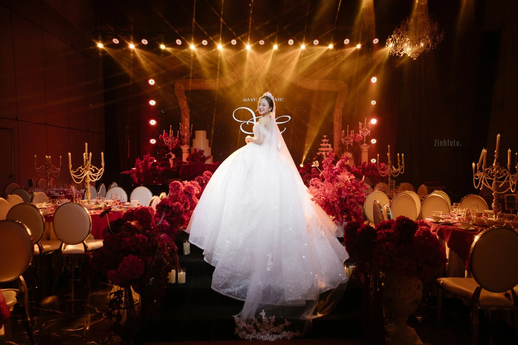 A bride in a white wedding gown smiling on a decorated wedding reception stage with pink flowers, candelabras, and a chandelier.