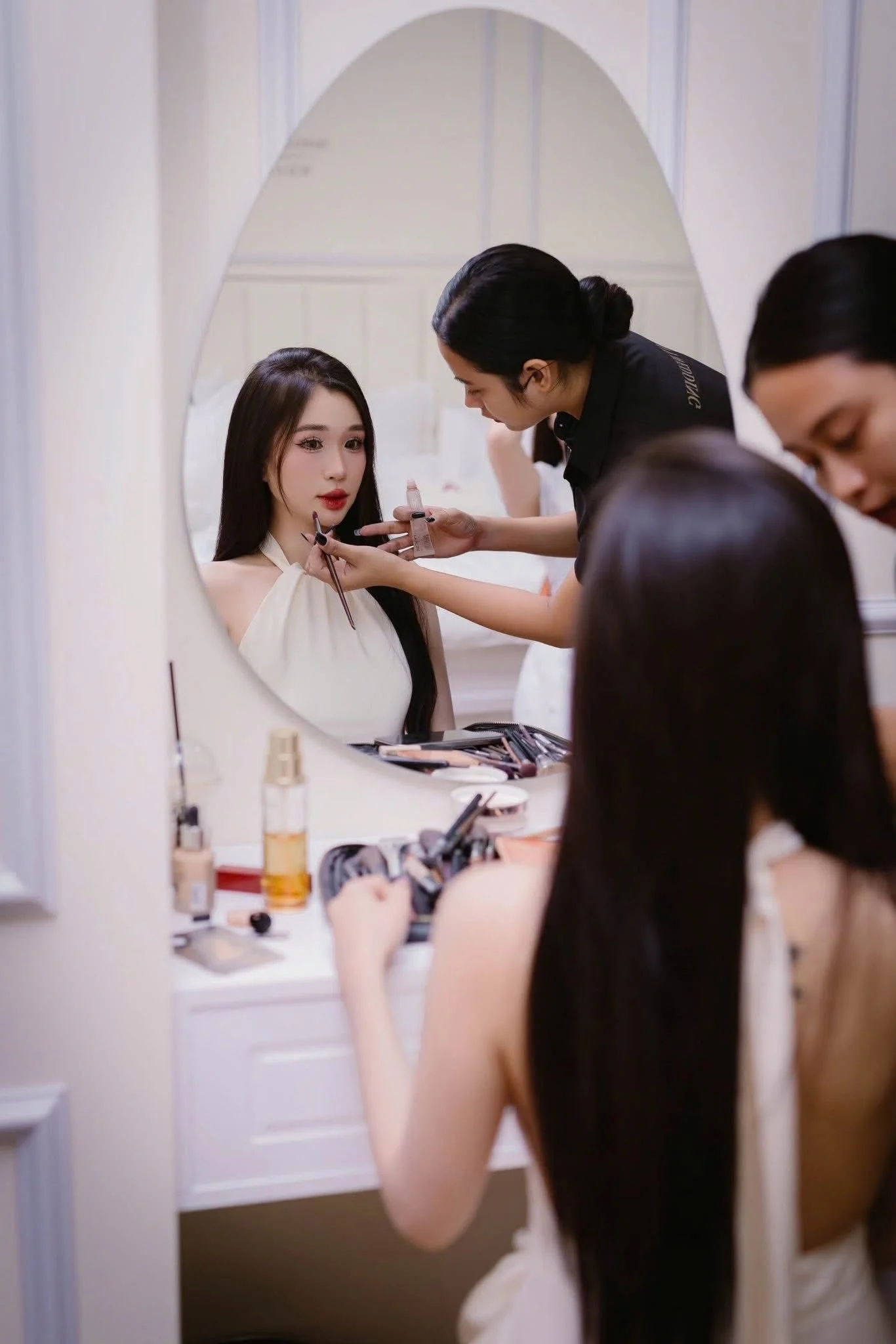A woman is having her makeup done by a makeup artist while sitting at a vanity table, reflected in an oval mirror. There are makeup products and brushes on the table, and another woman with long dark hair is sitting in front of her.