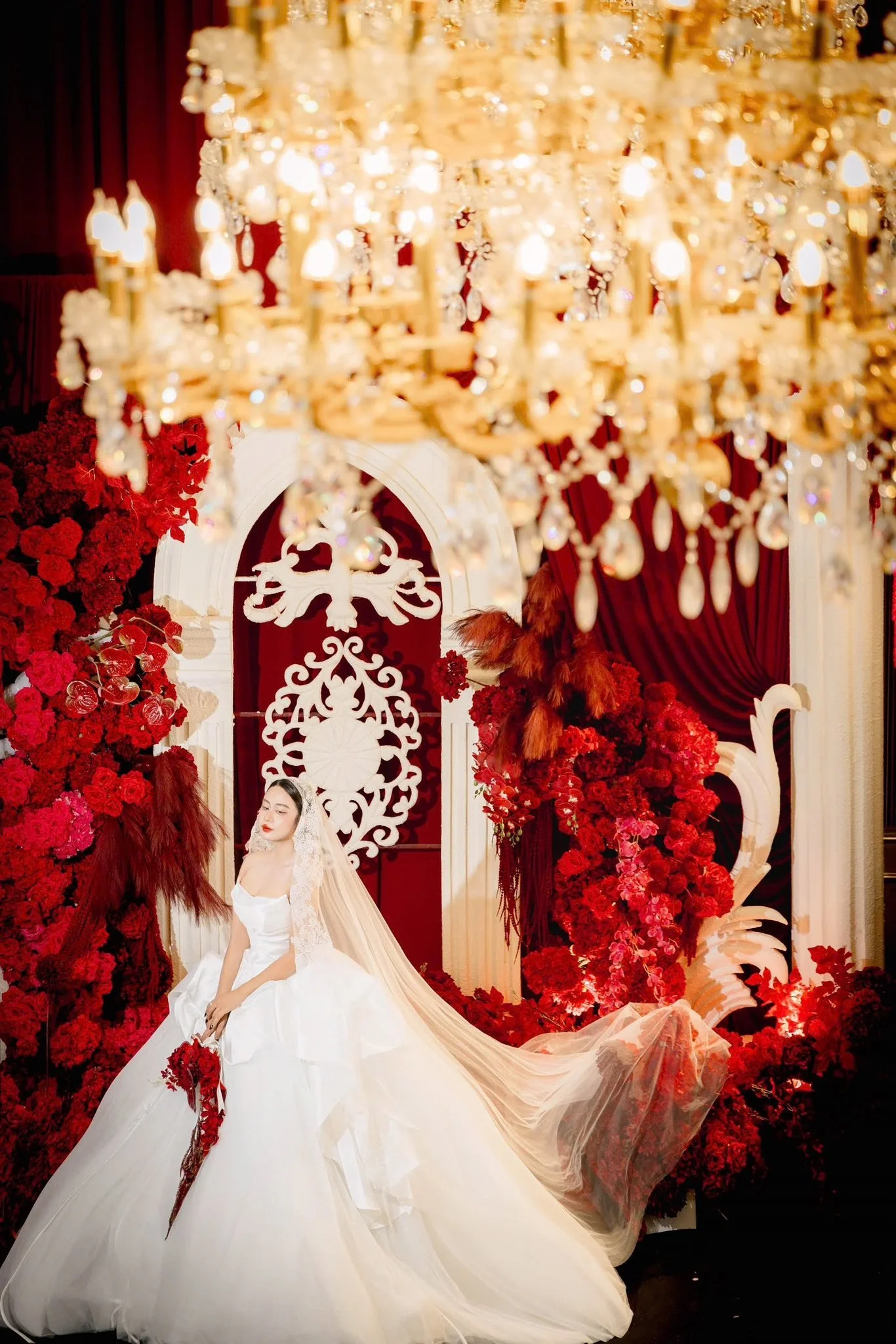 A bride in a white wedding gown with a long train, posing in an elaborate floral and curtain backdrop with a large chandelier above.