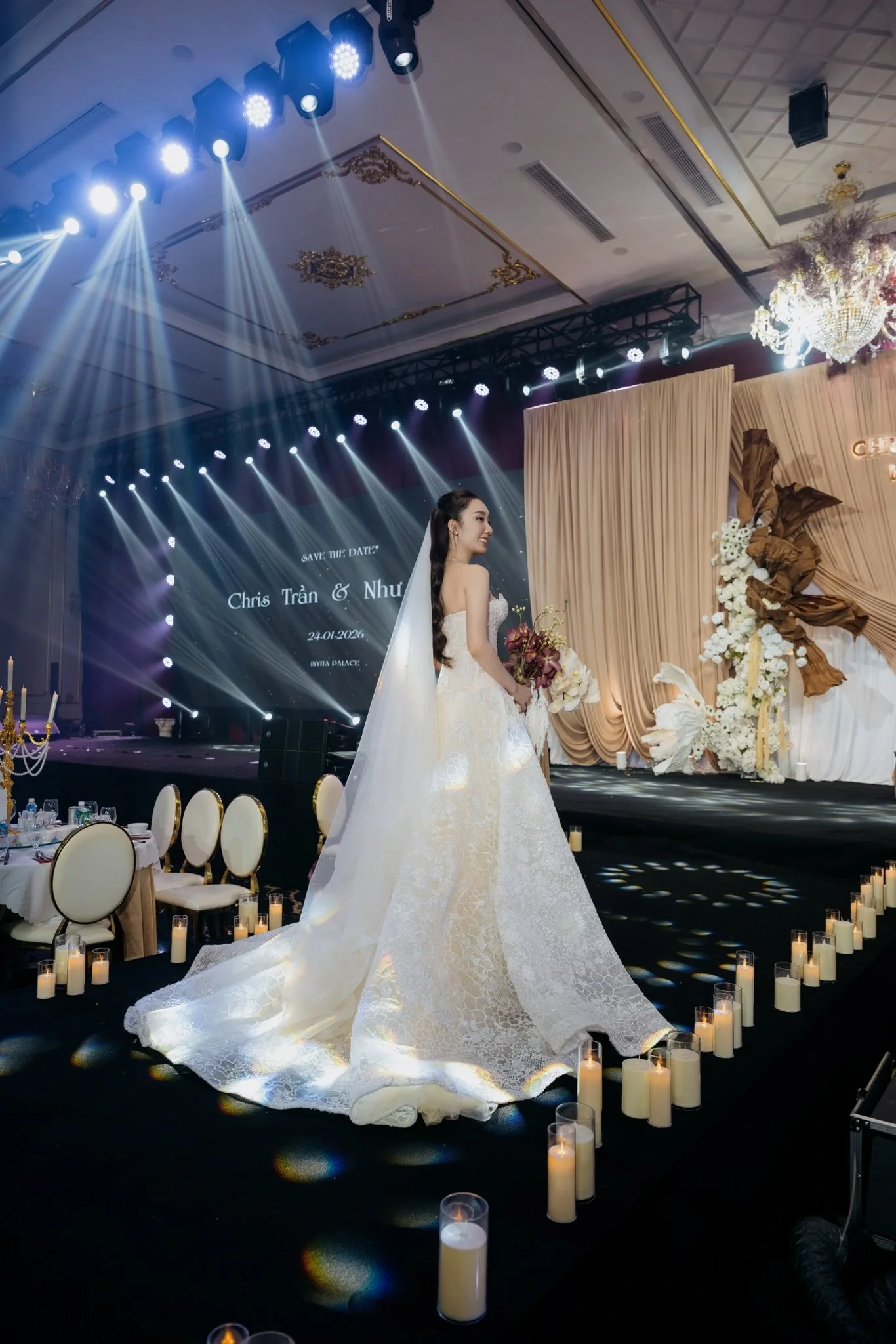 A bride in a white wedding gown stands on a stage holding a bouquet, amidst candles and elegant decor, with lighting and a backdrop that reads "Save the Date" and includes names Chris Trần and Nhu, and the date 24.01.2026.