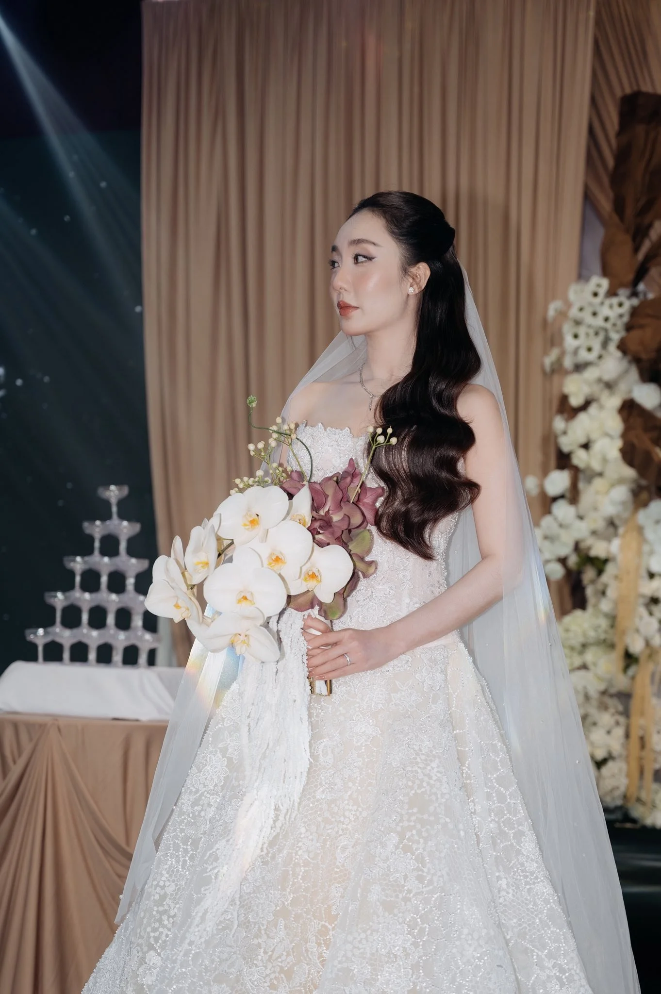 A bride holding a bouquet of white and purple orchids, wearing a white lace wedding gown and veil, standing in a decorated venue.