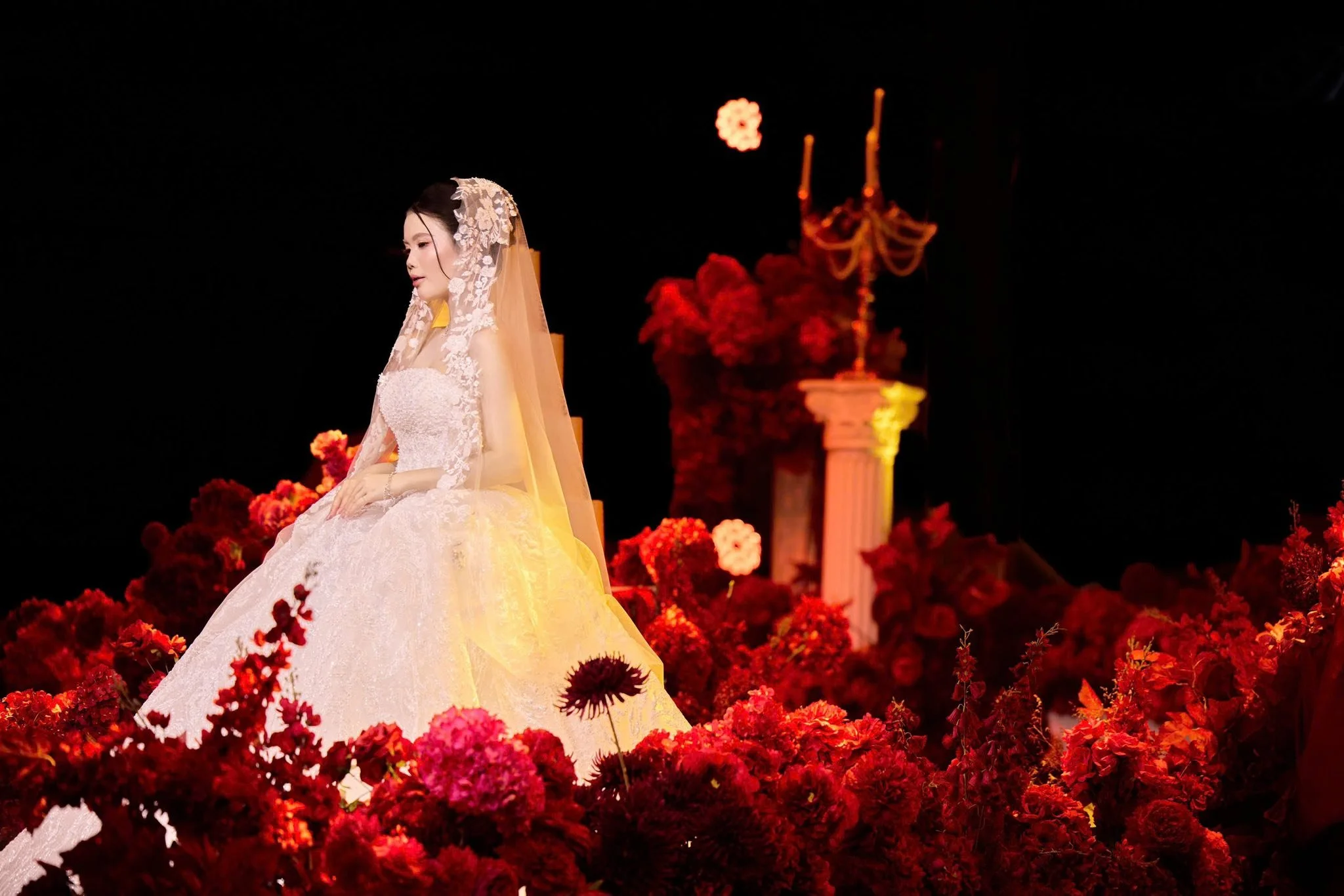 A bride in a white wedding dress and veil, sitting among red and pink flowers with decorative columns and candelabra in the background.