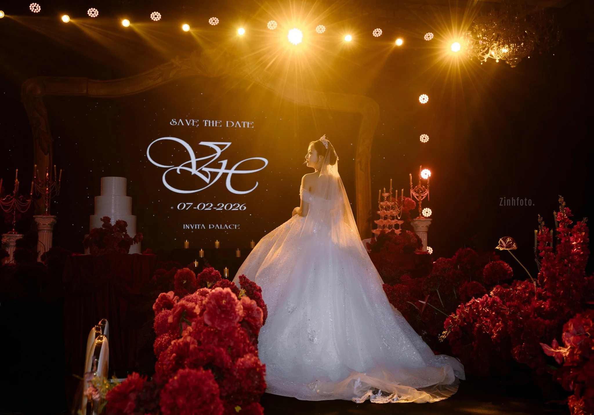 Bride in wedding gown on stage with 'Save the Date' for July 2, 2026, at Invita Palace, surrounded by red flowers and gold lighting.