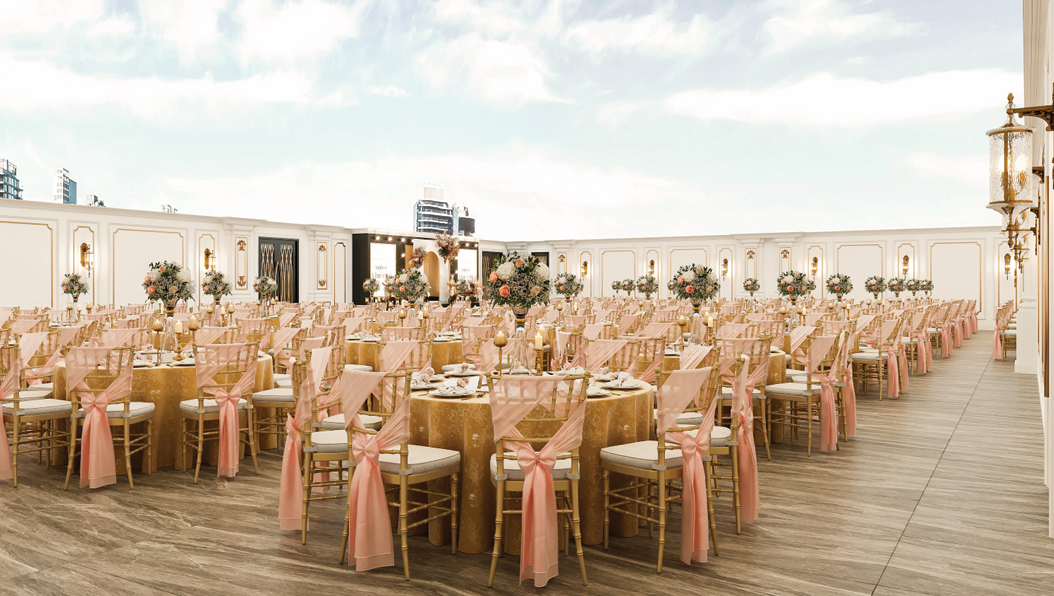 Elegant outdoor event space with numerous round tables covered in gold tablecloths, decorated with pink ribbon-wrapped chairs, floral centerpieces, and table settings, set against a white-paneled wall and a partly cloudy sky.