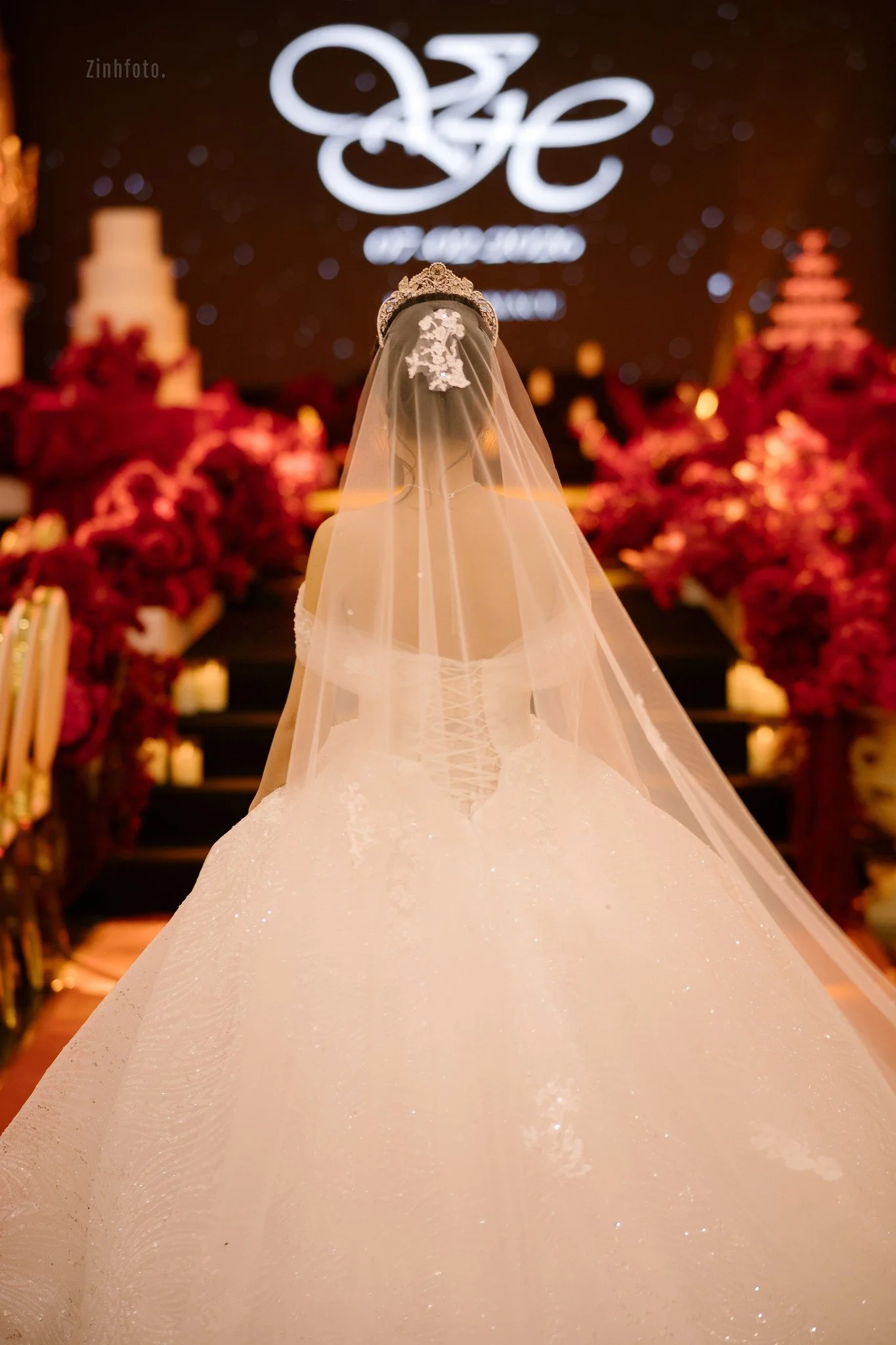Wedding dress with veil and tiara on mannequin, decorated with lace and sparkles, at a wedding venue with floral arrangements and a stage with a large screen in the background.