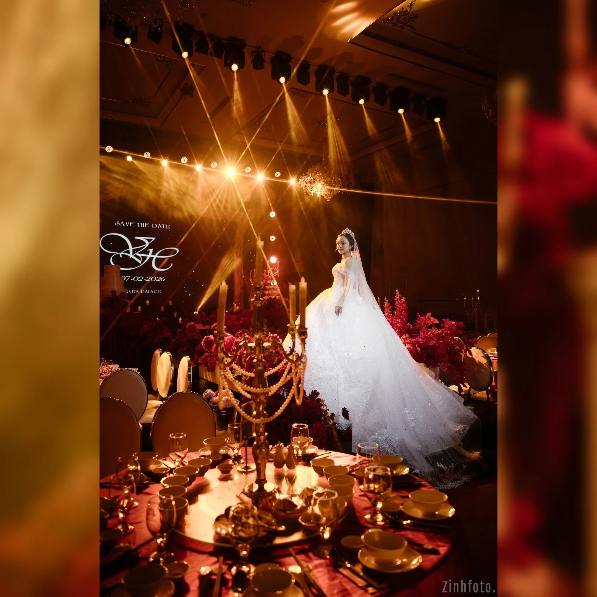 A bride in a wedding gown standing on stage surrounded by flowers and decorative candles, illuminated by warm stage lights at a wedding reception.