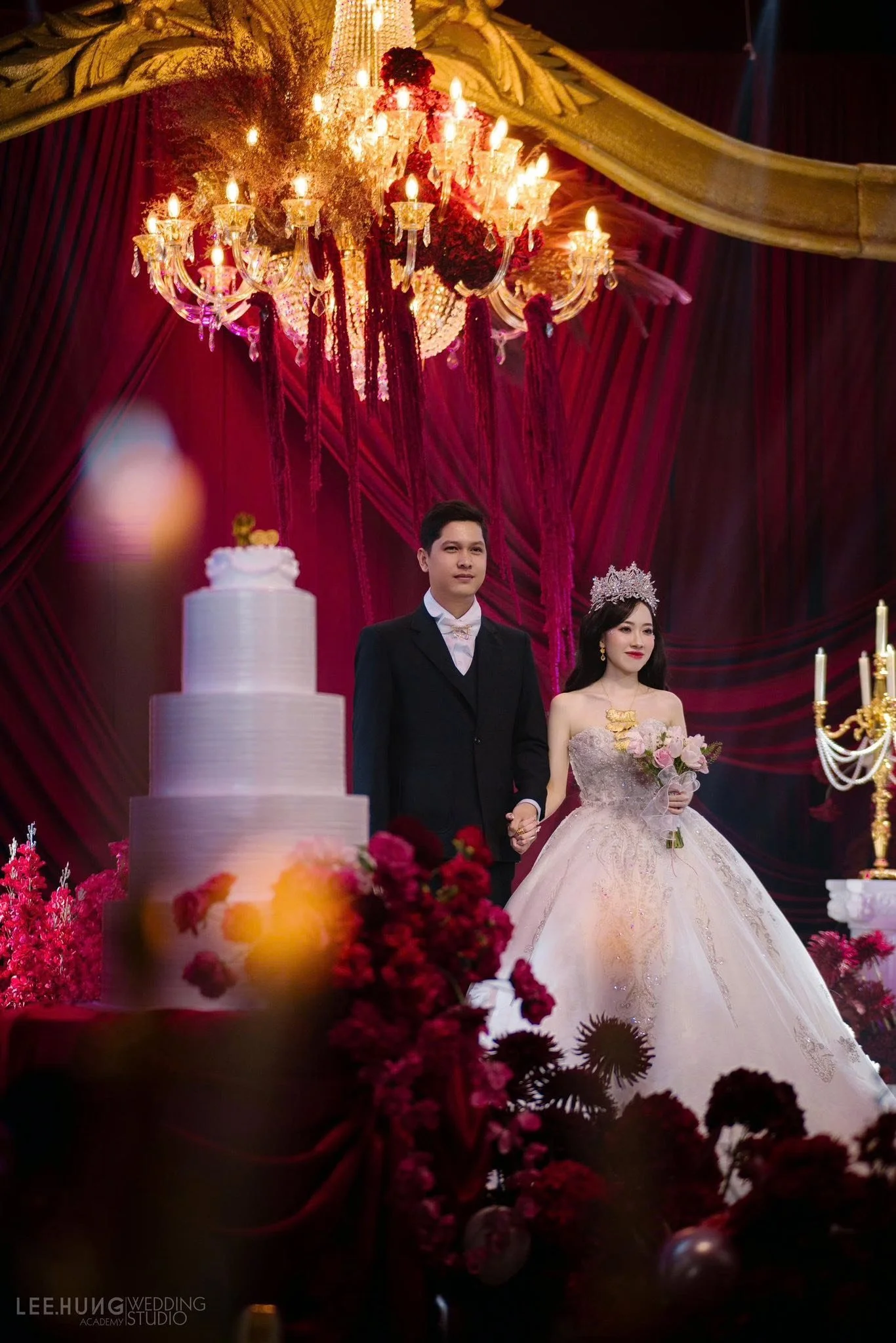 A bride and groom standing together at their wedding reception, decorated with flowers, a large tiered wedding cake, and ornate chandeliers, with a red velvet background.